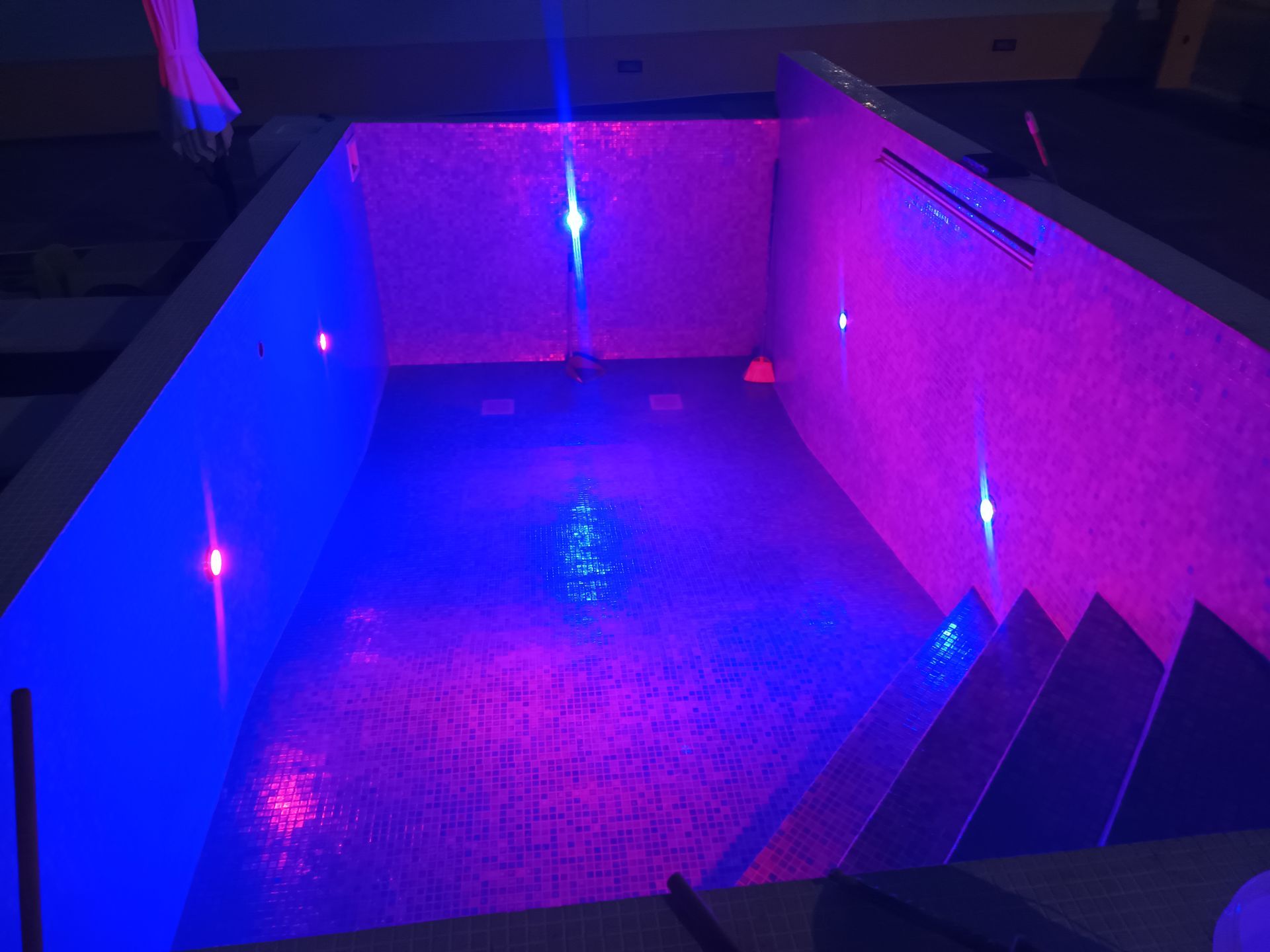 Pequena piscina coberta com luzes azuis e roxas, degraus e paredes texturizadas.