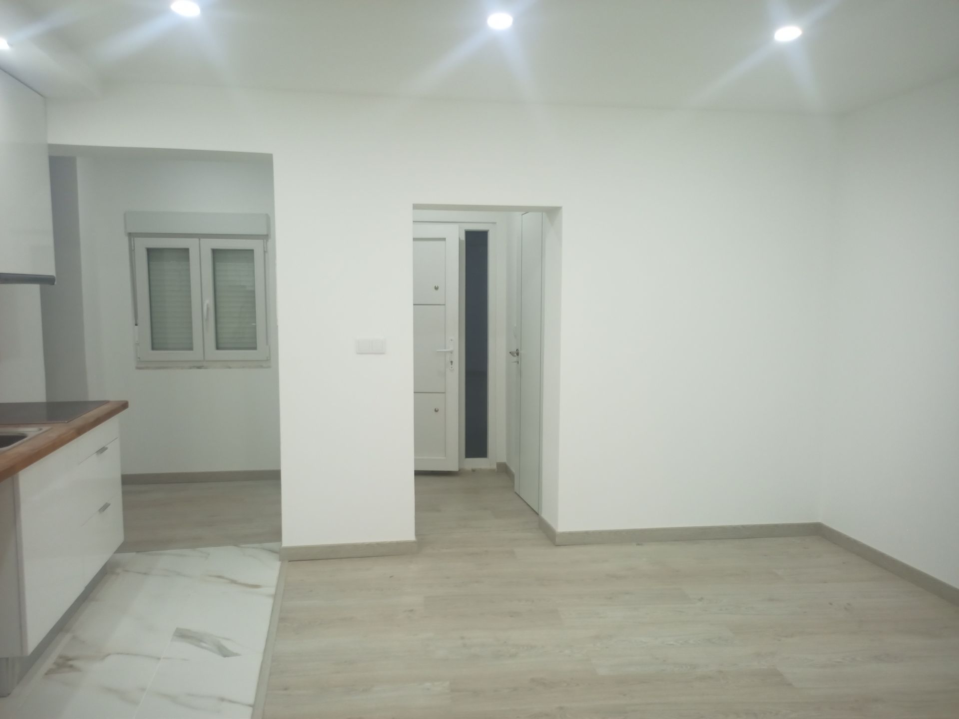 Vista interior de um cômodo predominantemente branco com portas que dão acesso à cozinha e a outro cômodo.