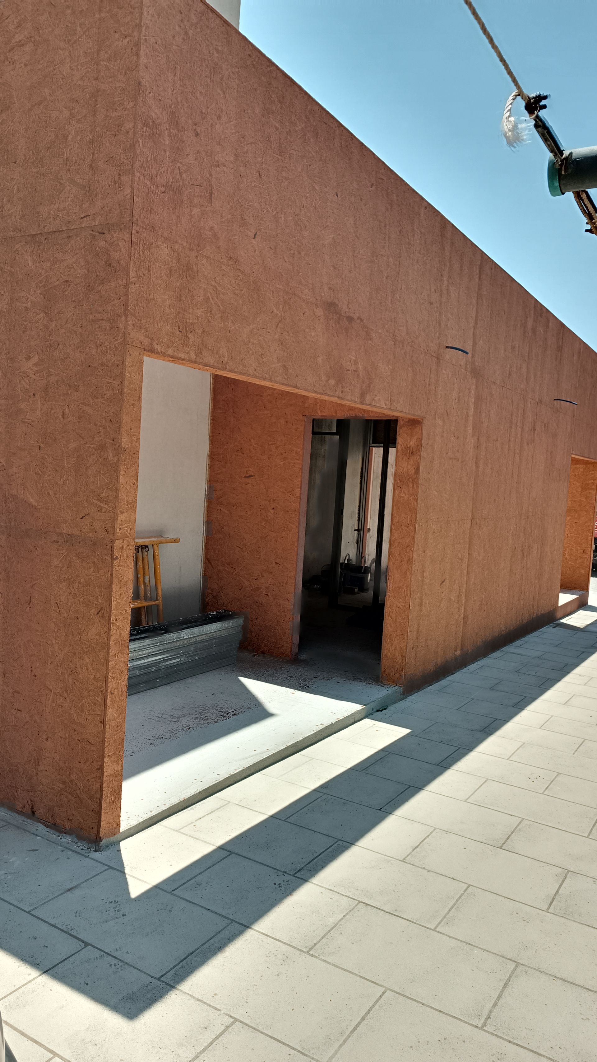 Vista exterior de um edifício com uma abertura na entrada, feita de painéis de madeira castanho-avermelhada, num dia ensolarado.