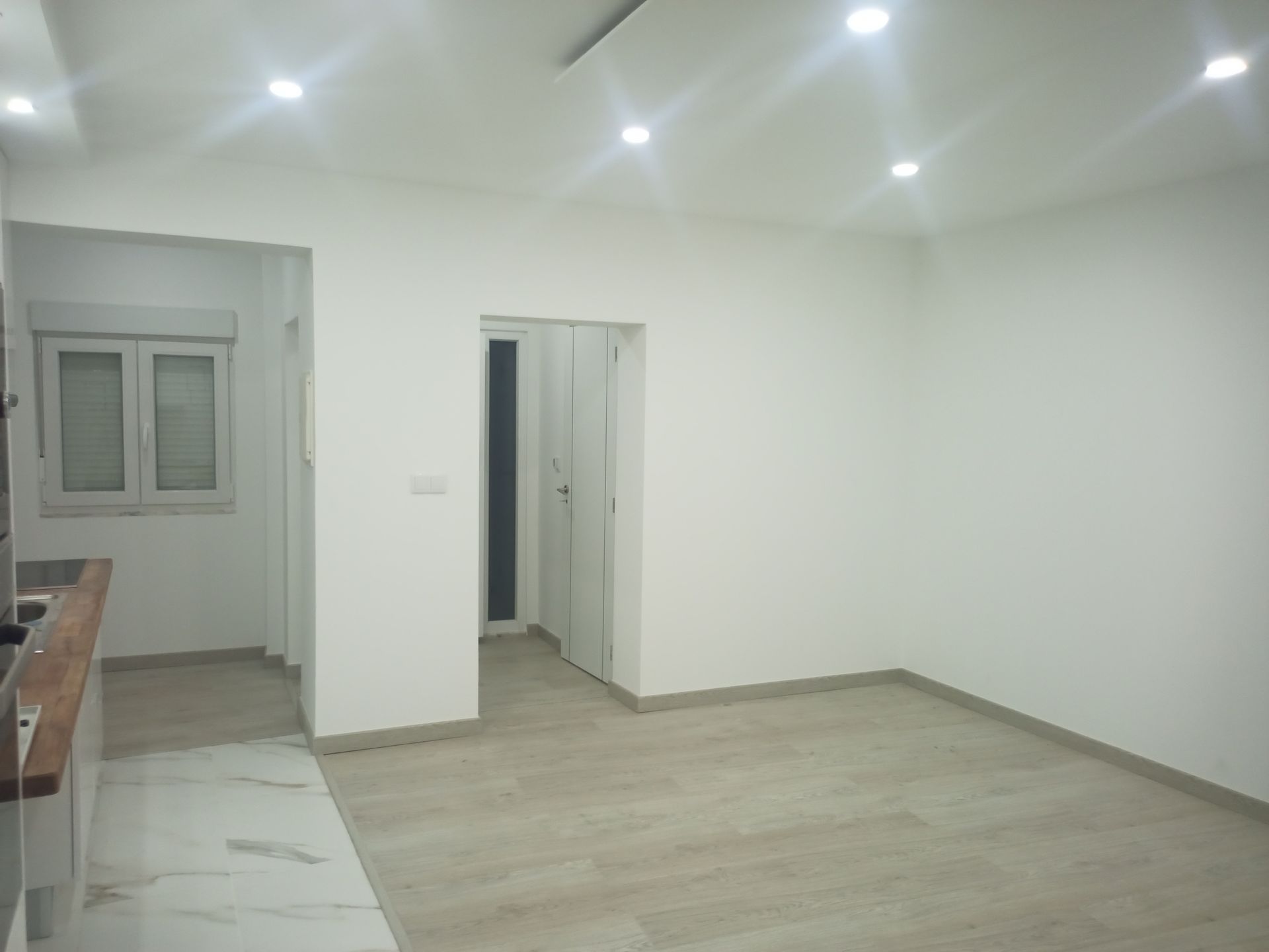 Sala vazia com paredes brancas, iluminação embutida e piso neutro; porta para o corredor e janela.