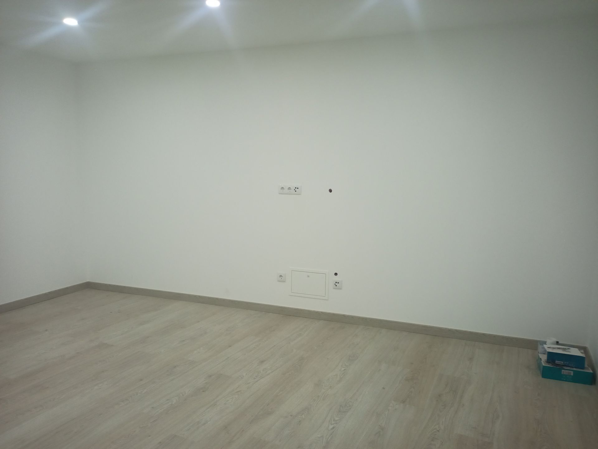 Quarto vazio com paredes brancas, piso de madeira clara e tomadas elétricas.