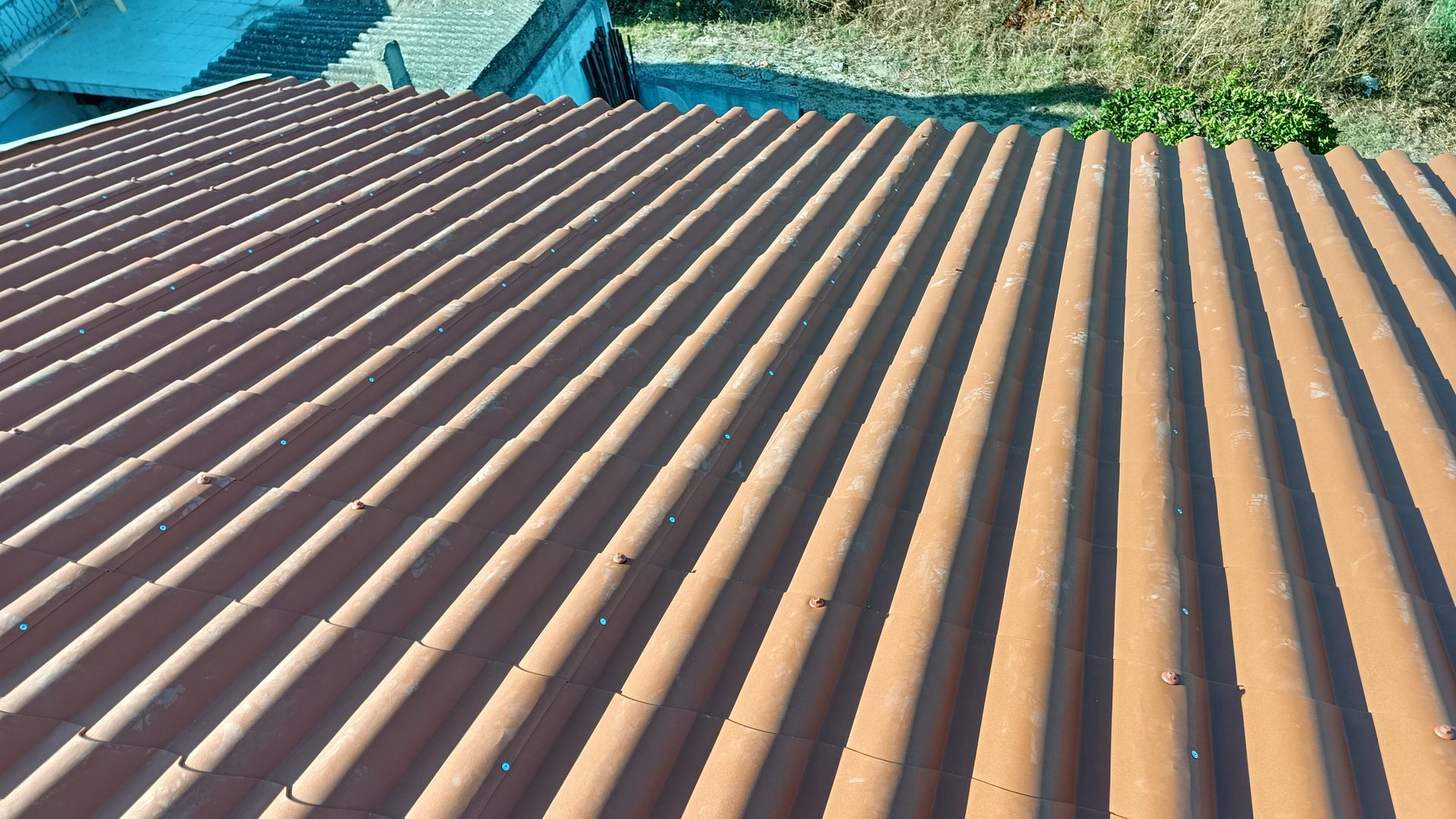 Telhado marrom ondulado banhado de sol, com vista parcial para a área verde adjacente.