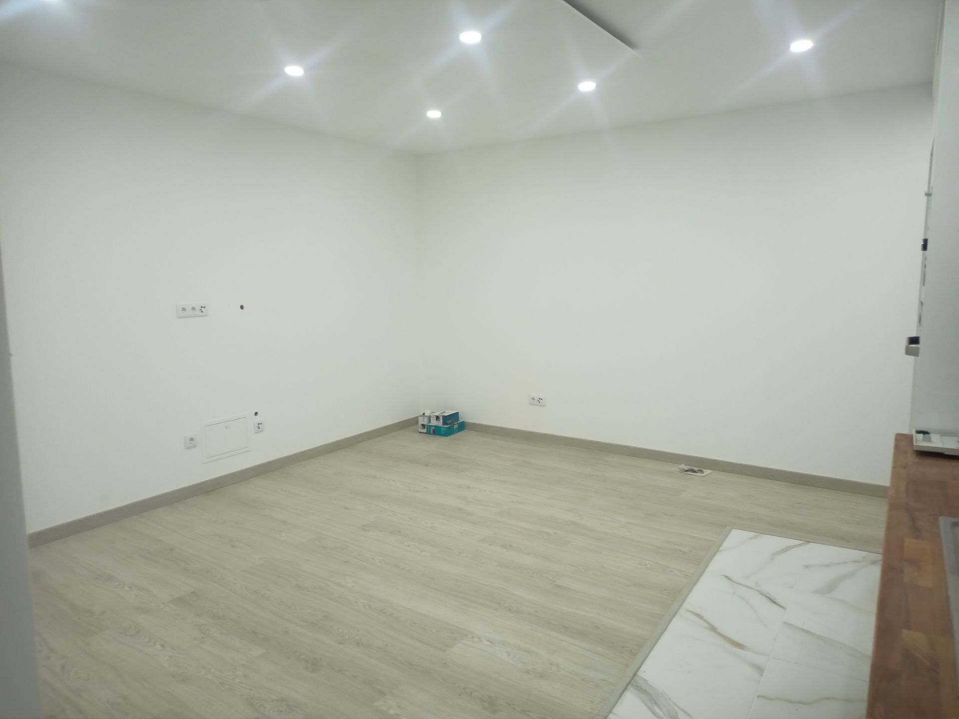 Quarto vazio com paredes brancas, piso que imita madeira clara e luzes embutidas no teto.