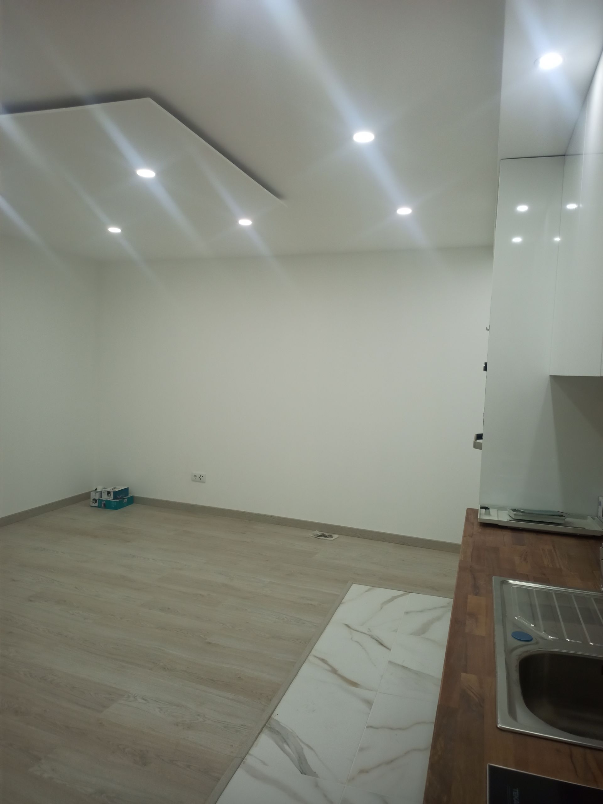 Sala vazia com piso imitando madeira, paredes brancas, iluminação embutida no teto e bancada de cozinha com pia.