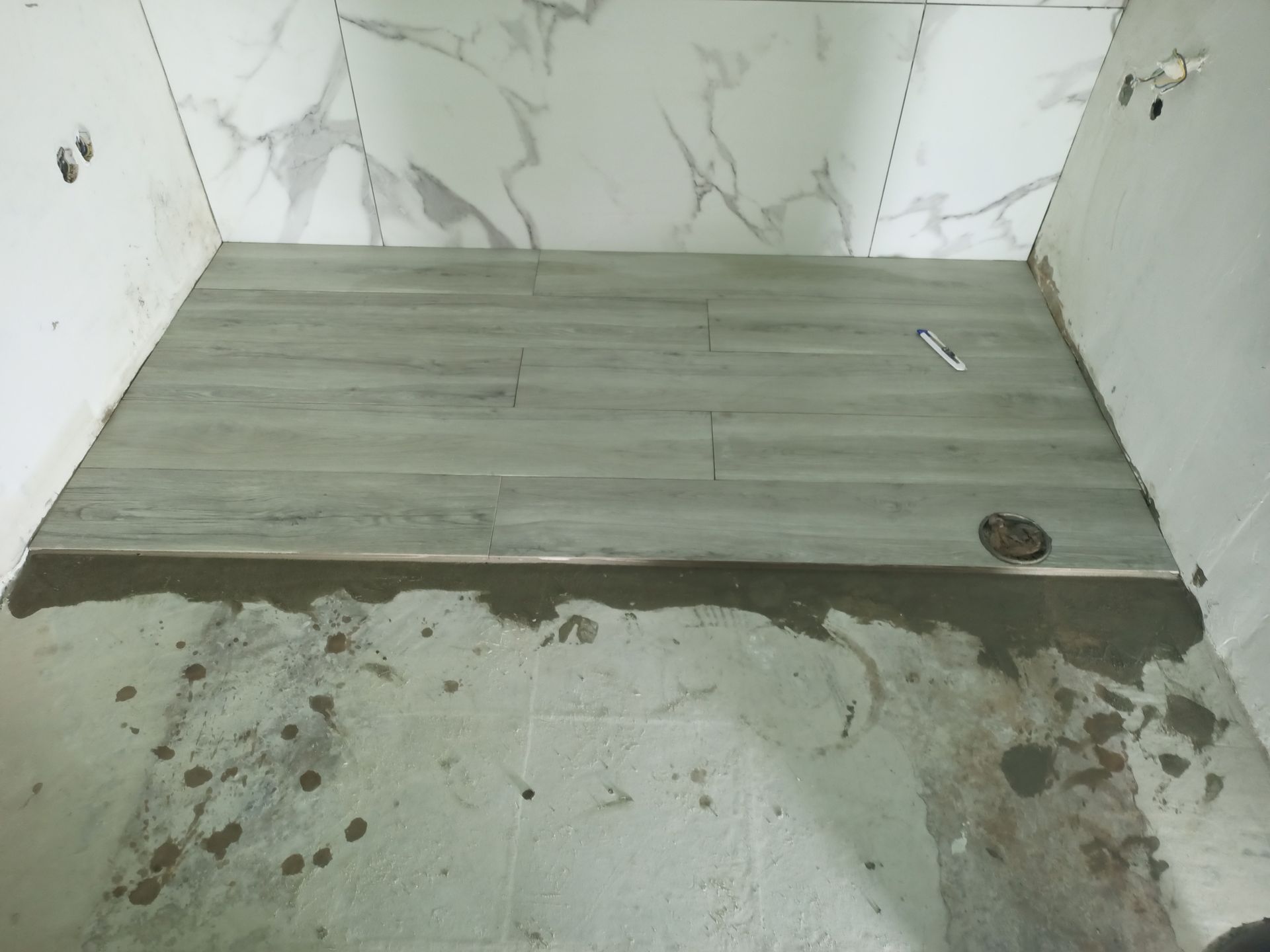 Instalação de azulejos no piso do banheiro em andamento: azulejos cinza imitando madeira, parede superior com azulejos com estampa de mármore.