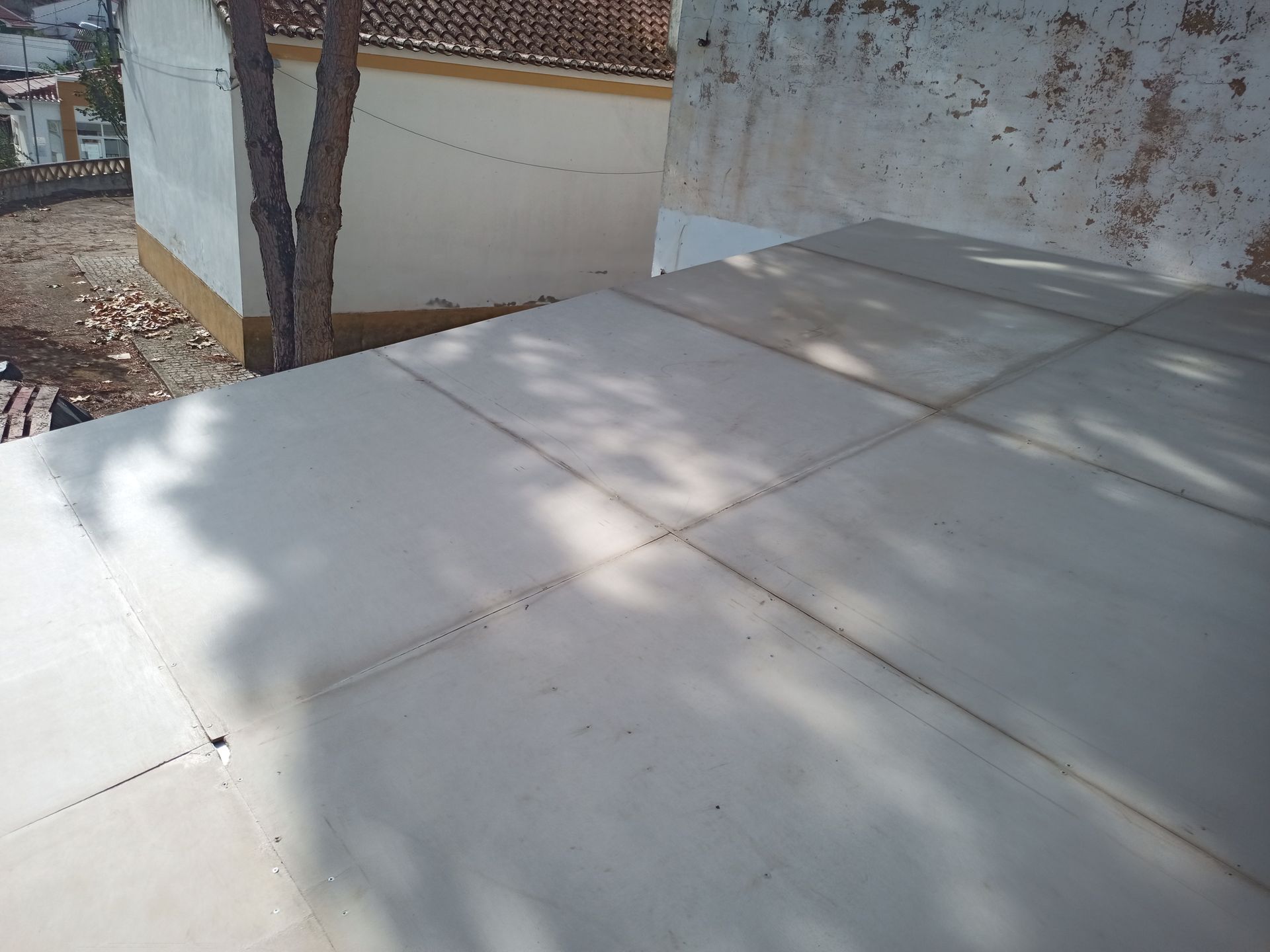 Telhado de telhas cinzentas com sombras, adjacente a uma parede branca desgastada e outras estruturas.