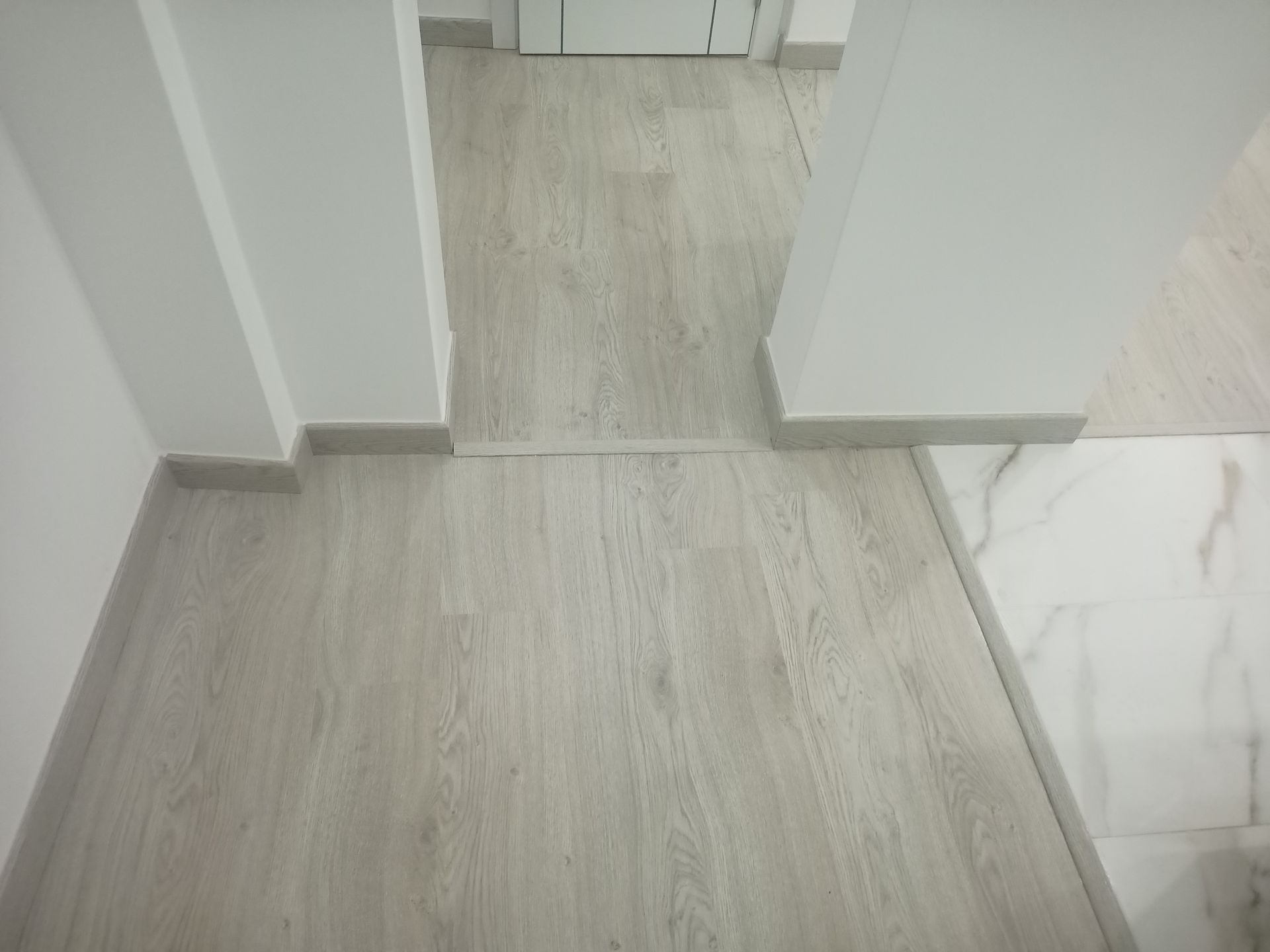 Piso com aparência de madeira clara e fresta visível, delimitado por paredes brancas e rodapés cinza.