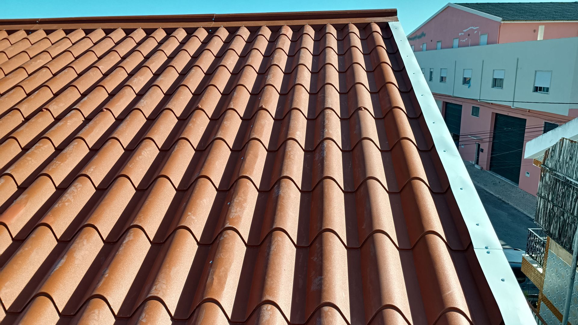 Telhado de telha de barro marrom com borda de metal branco, prédios ao fundo.