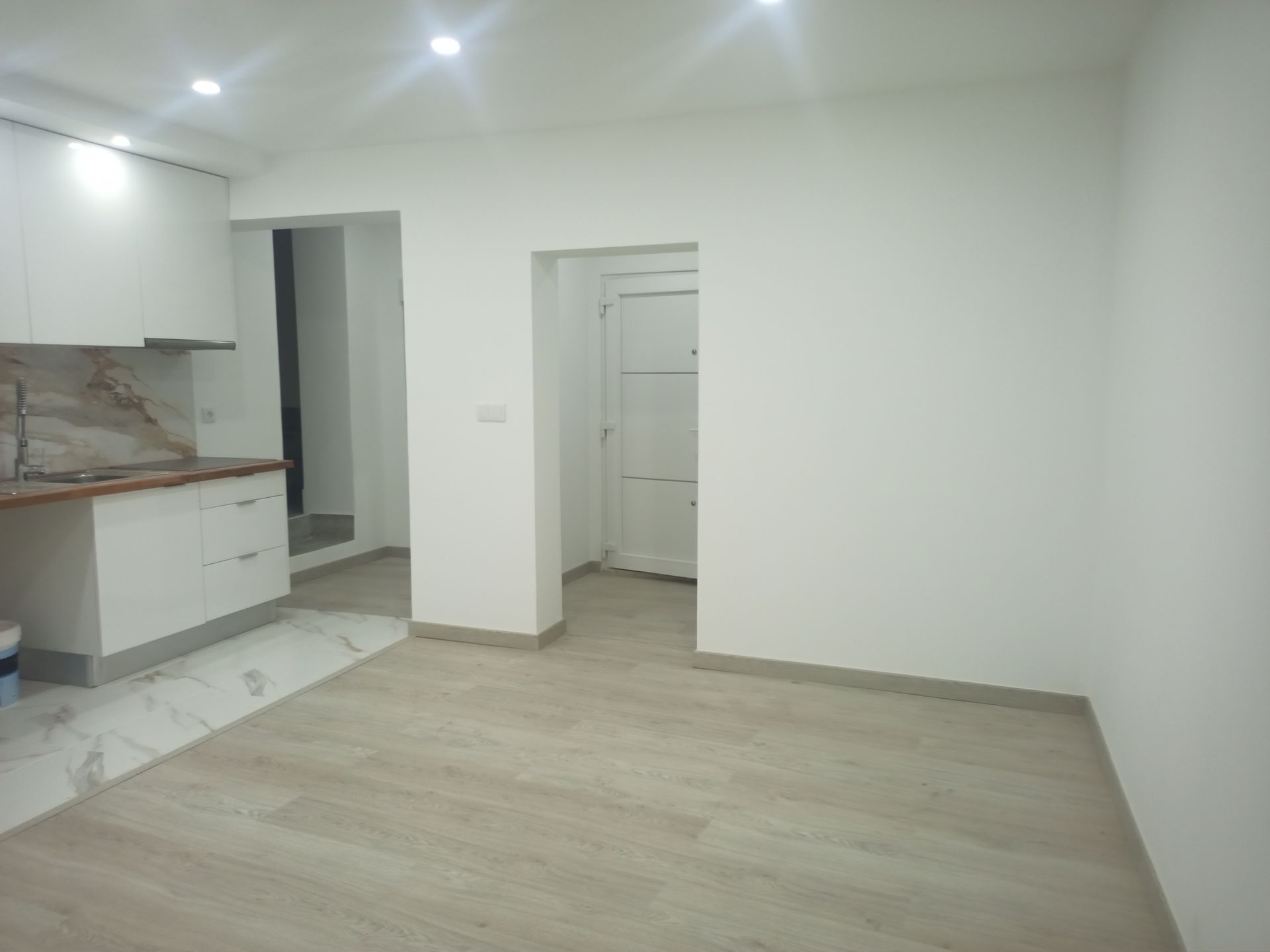 Sala vazia com área de cozinha; paredes brancas, piso que imita madeira clara.