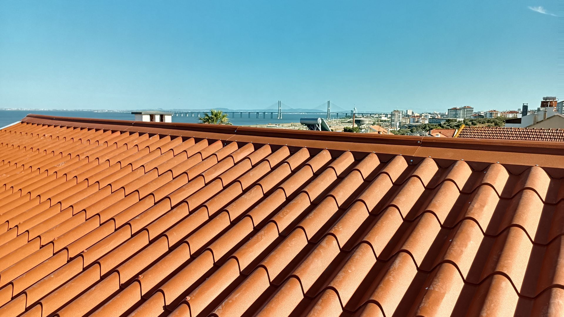 Telhado de telha vermelha com uma cidade costeira e uma ponte ao fundo, sob um céu azul.