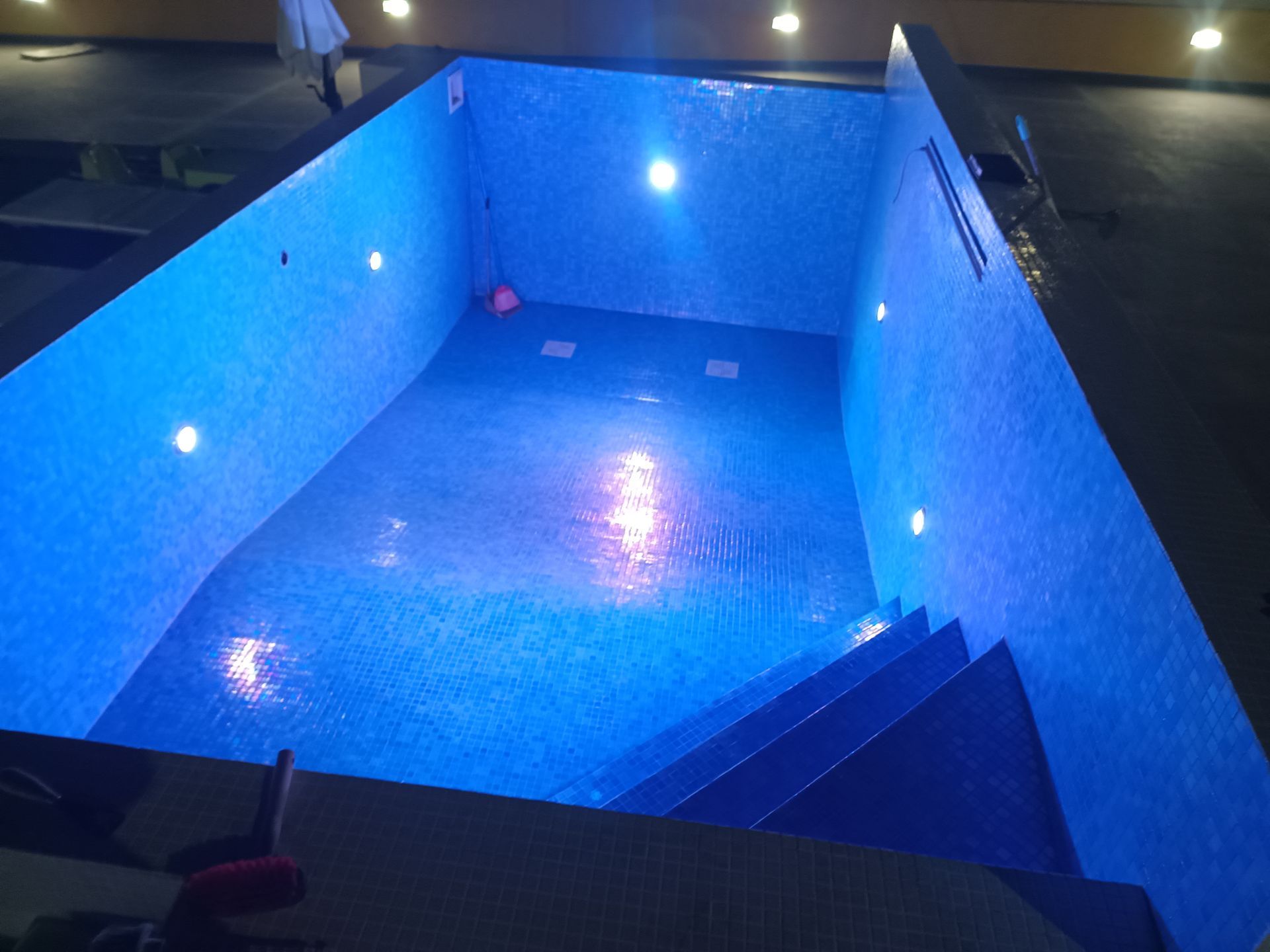 Piscina retangular revestida com azulejos azuis e iluminada com luzes azuis. Degraus descem até a água.