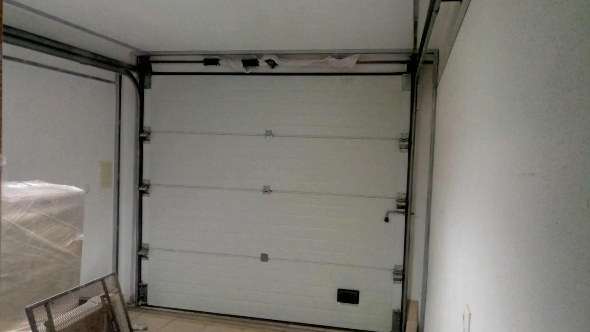 Porta de garagem branca fechada com moldura de metal, em uma sala branca vazia.