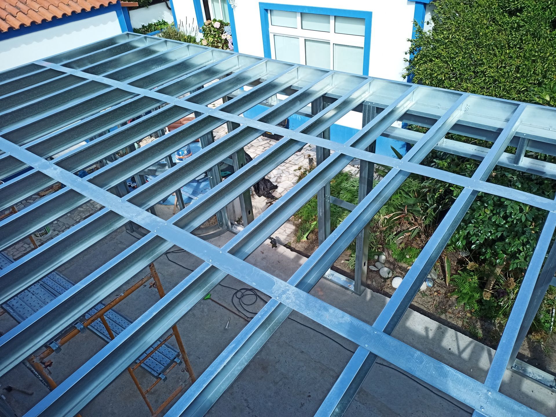 Estrutura de cobertura em metal, parcialmente construída sobre uma superfície de concreto, com vista para um edifício e área verde.