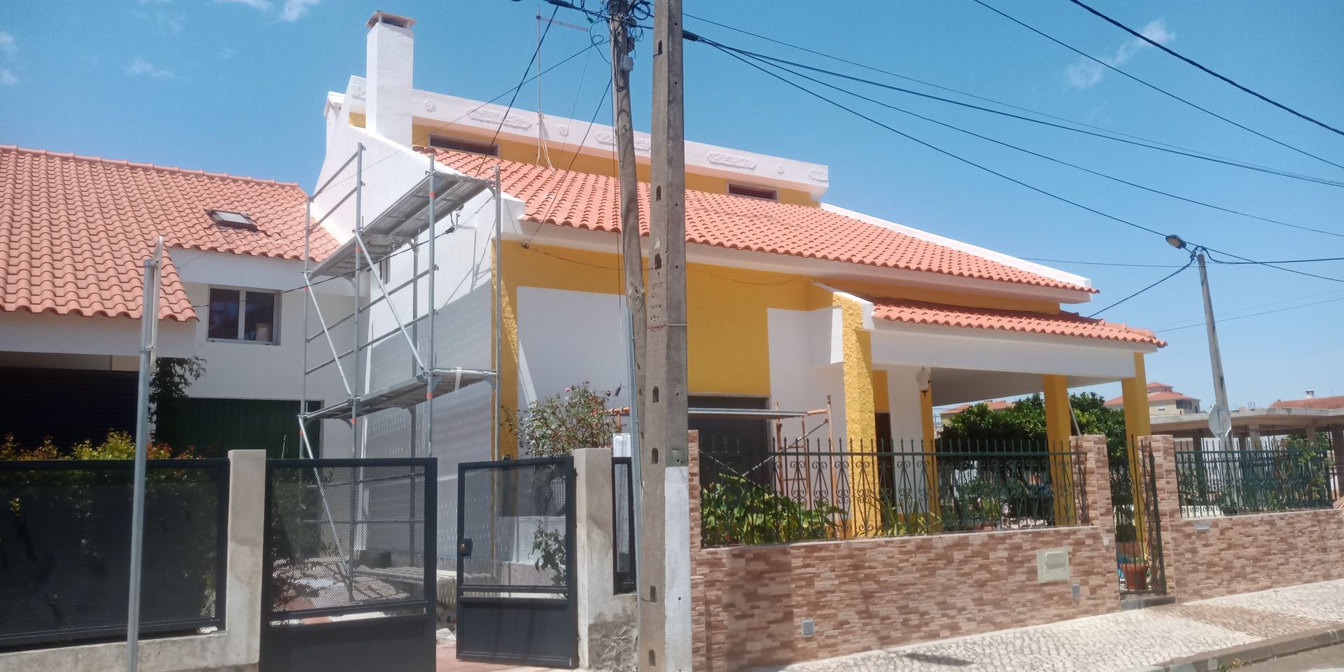 Uma casa de dois andares com telhado de telhas vermelhas, paredes amarelas e chaminé branca está localizada atrás de um muro de tijolos e um portão preto.