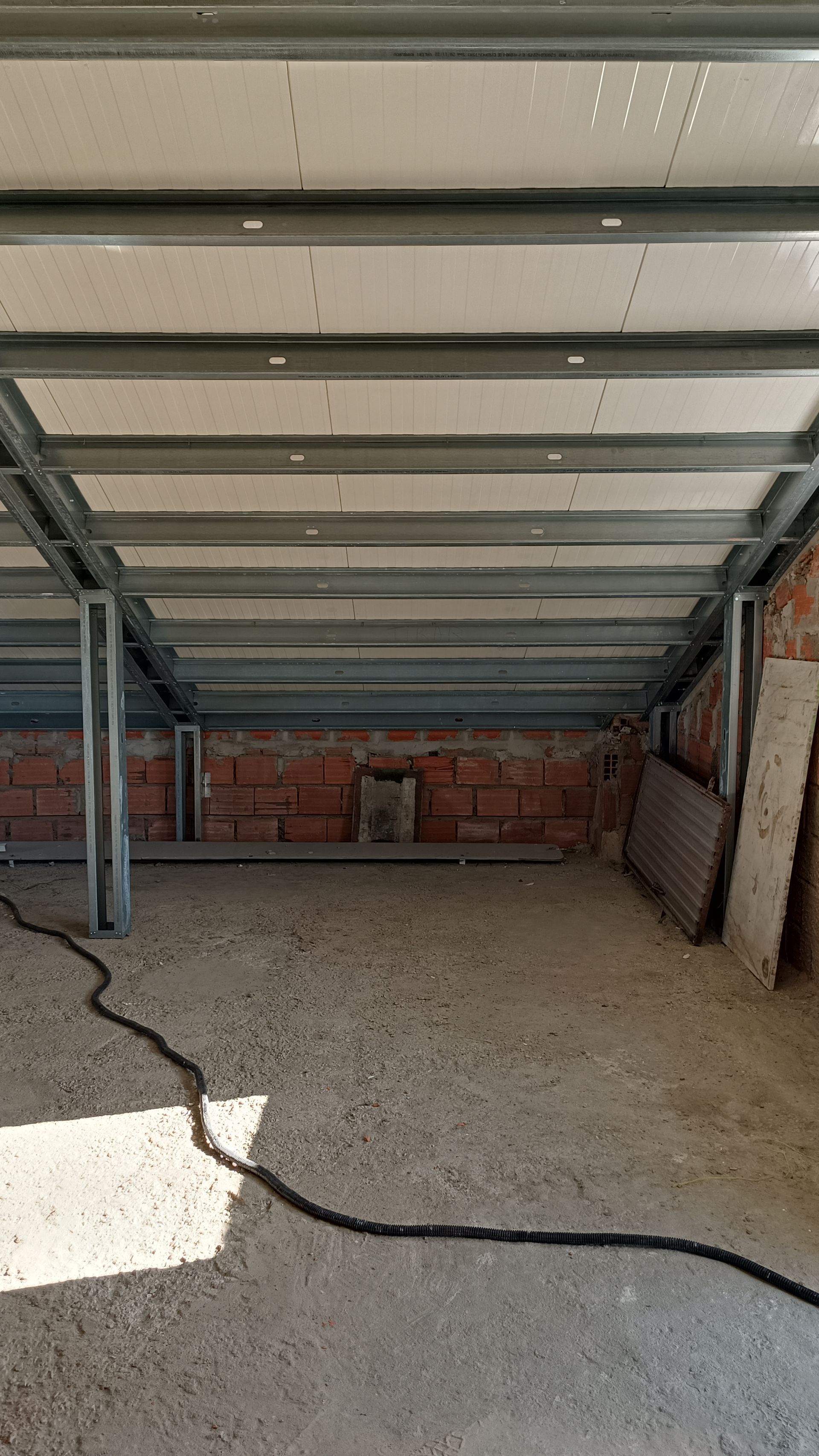 Canteiro de obras: teto com estrutura metálica e painéis brancos, parede de tijolos aparentes, piso de concreto, cabeamento preto.