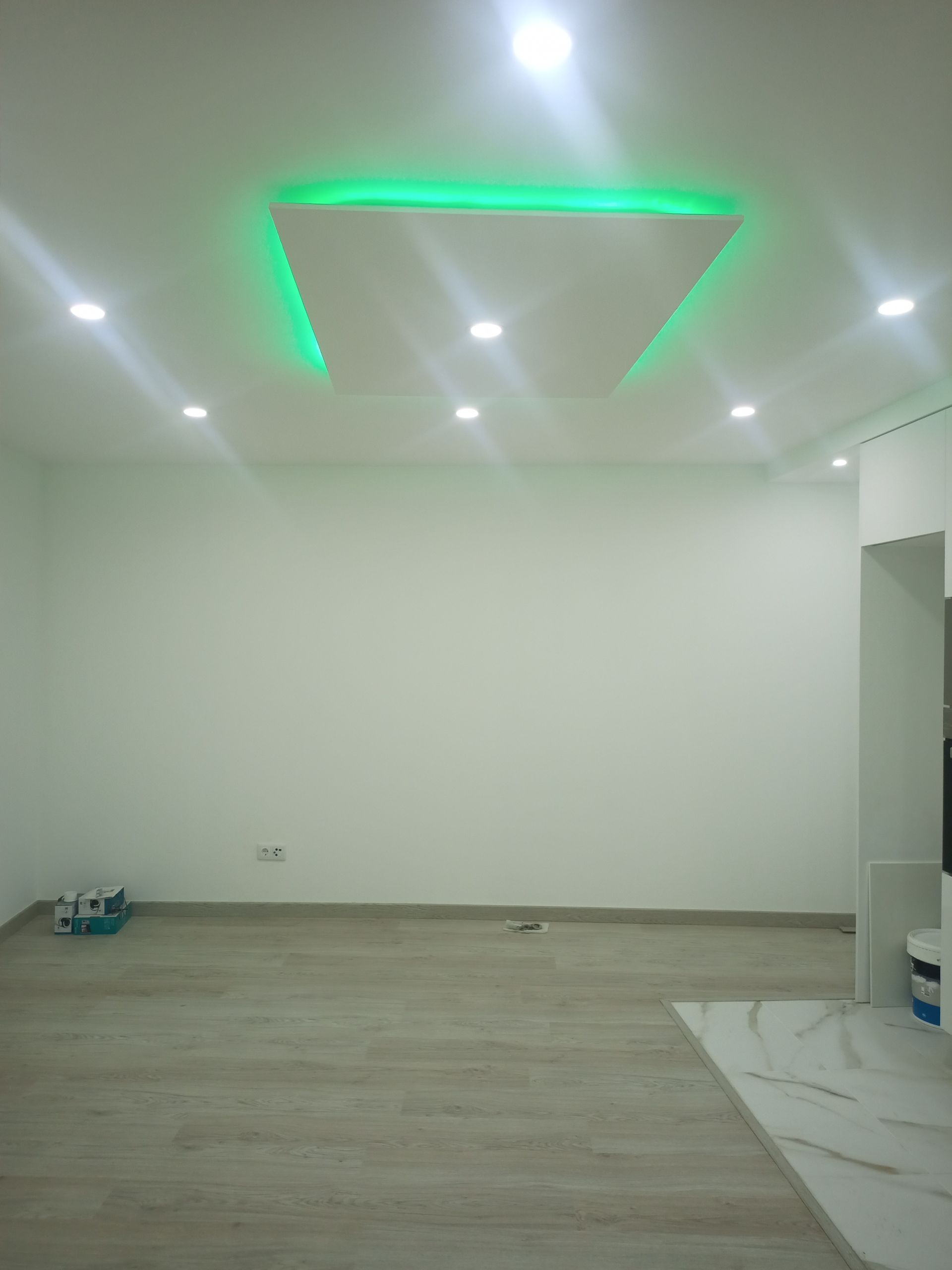 Quarto vazio com paredes brancas, luzes embutidas no teto e detalhes em verde. Piso de madeira clara.