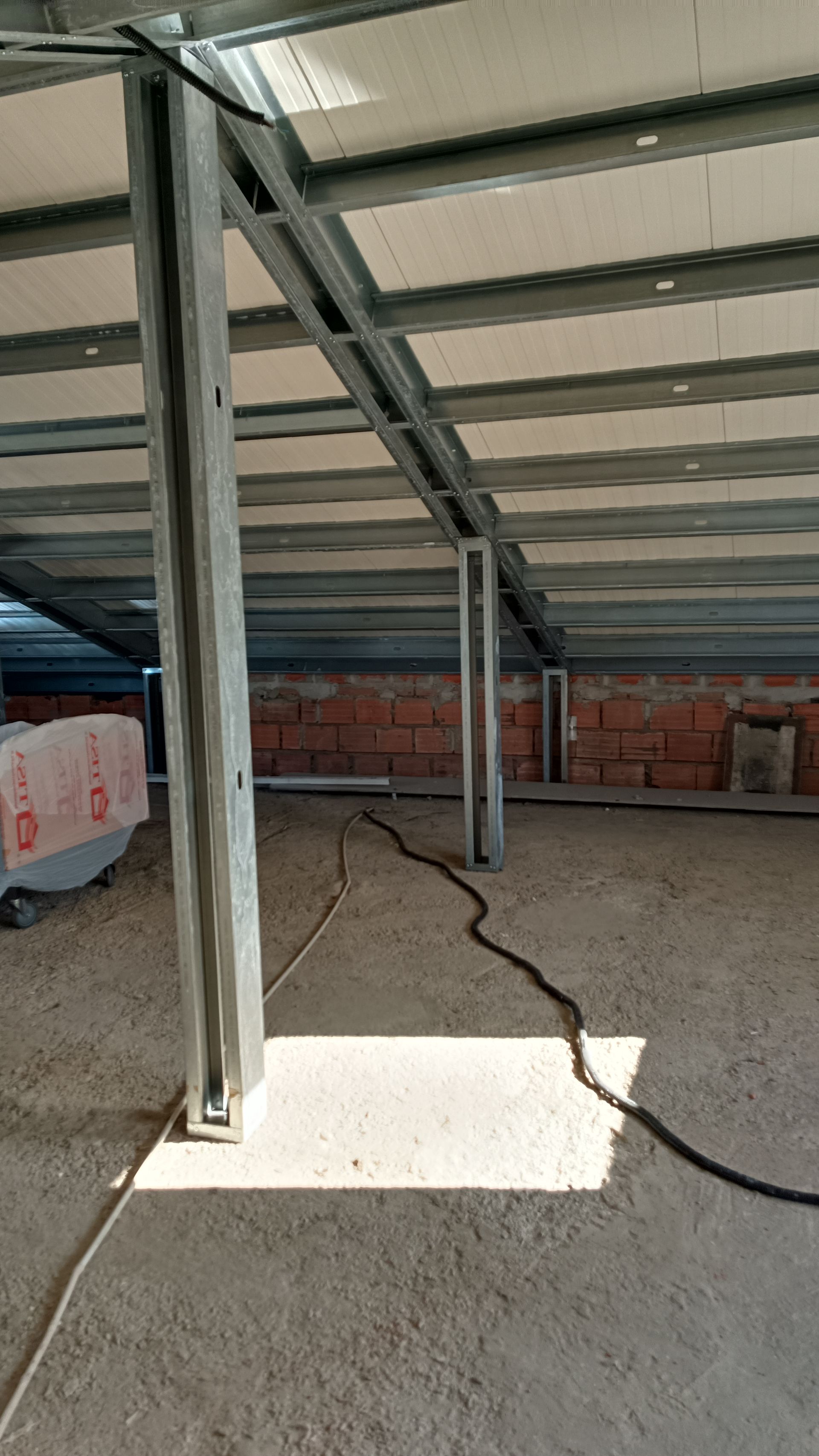 Vista interna de um canteiro de obras com estruturas de aço, piso de concreto e parede de tijolos ao fundo.