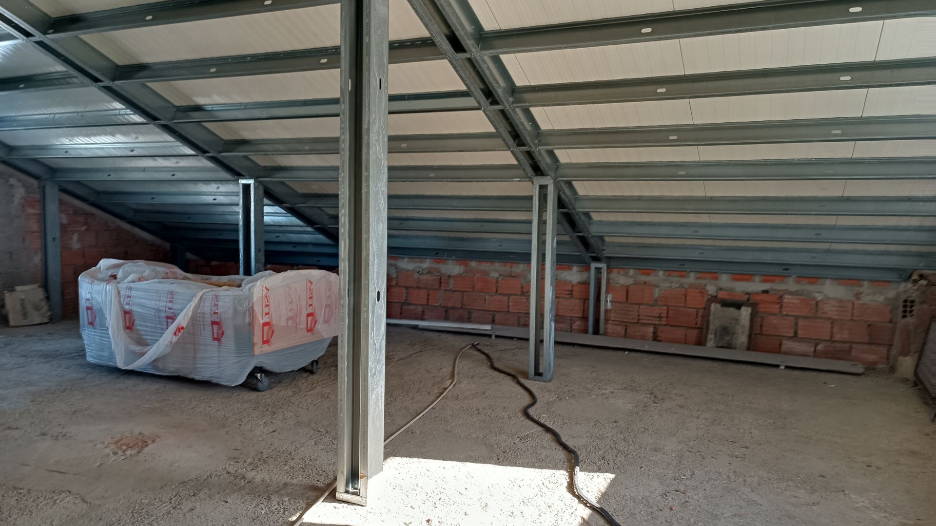 Construção interna de um cômodo, mostrando estrutura metálica, parede de tijolos e piso de concreto.