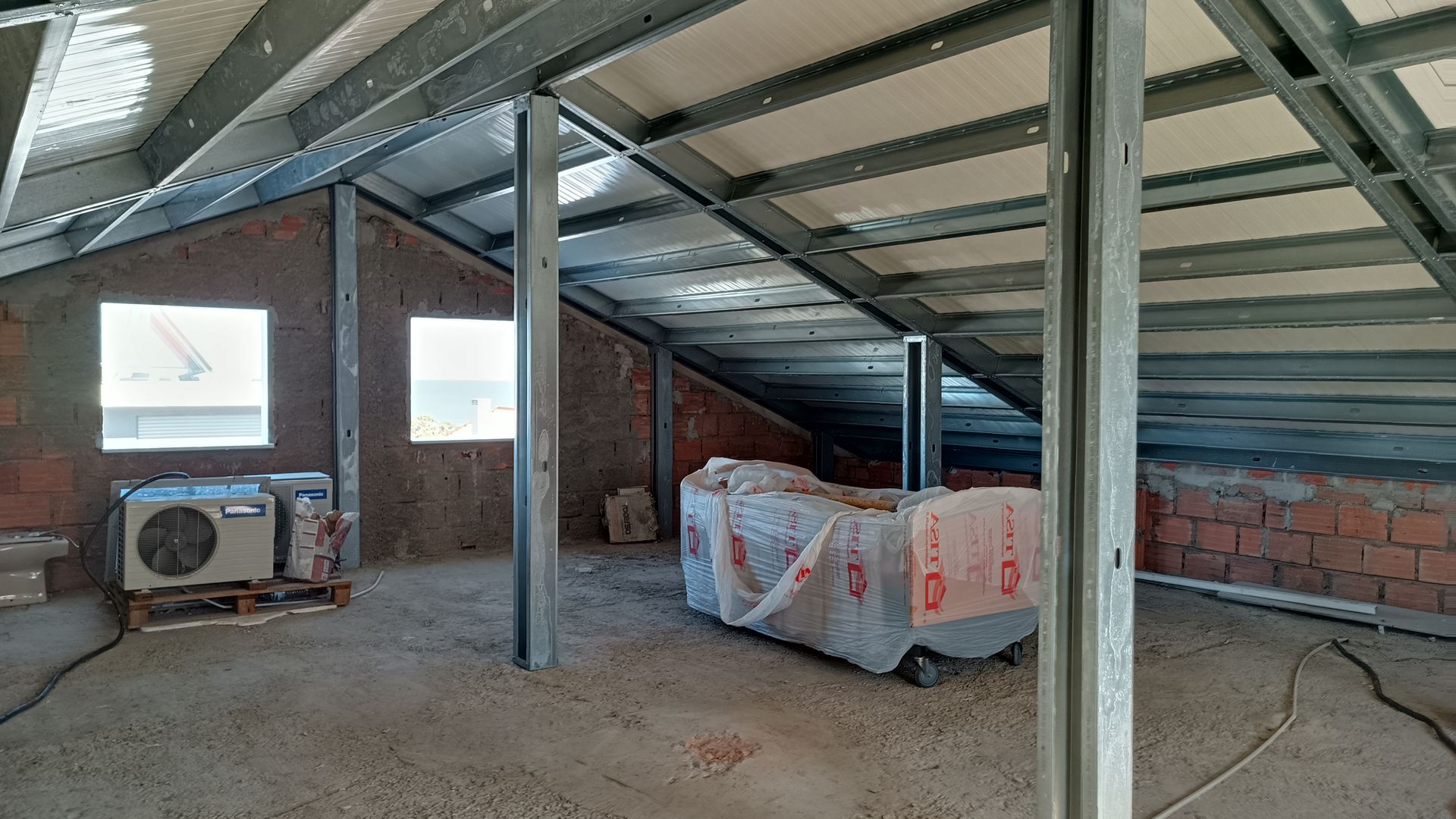 Interior de um sótão inacabado com estrutura metálica exposta, paredes de tijolo e duas janelas.