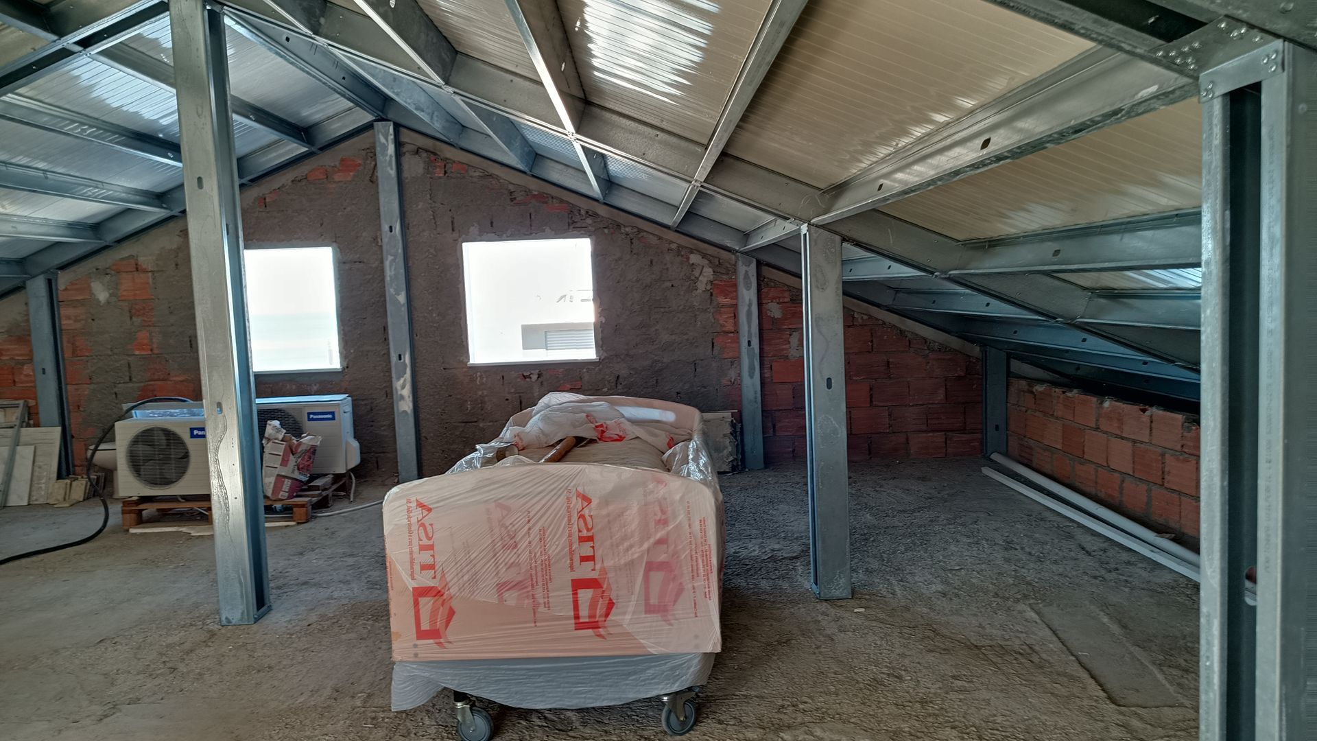 Interior de um canteiro de obras com estrutura metálica exposta, paredes de tijolos e duas pequenas janelas.