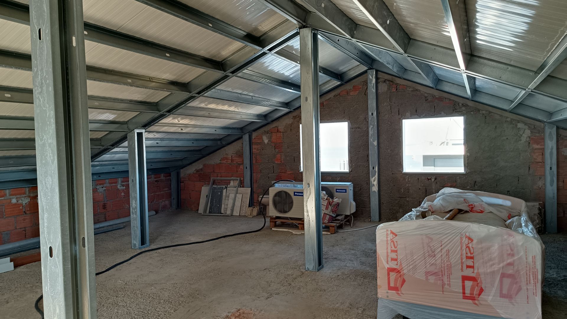 Interior de um cômodo em construção com vigas de metal, paredes de tijolos aparentes e duas pequenas janelas.