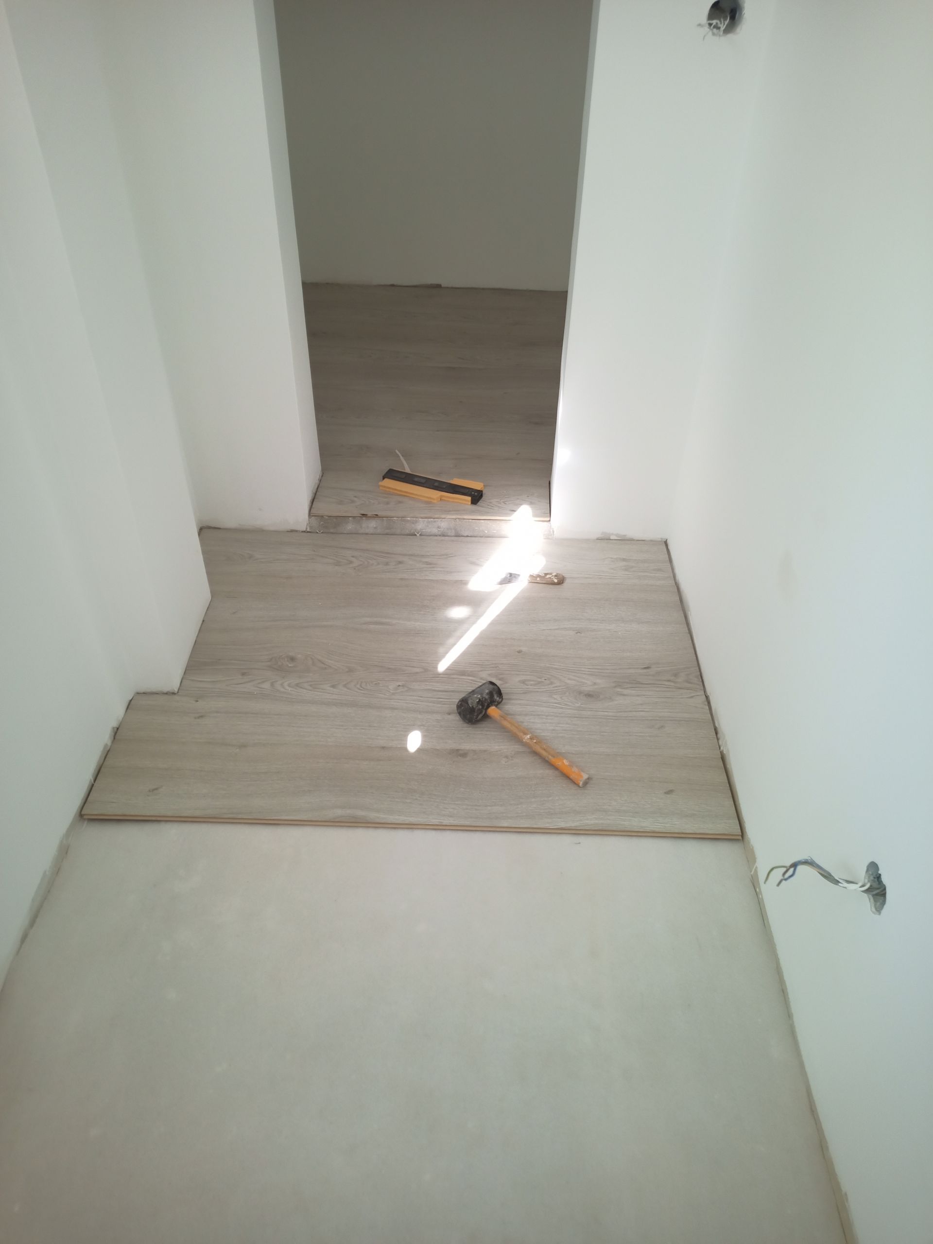 Instalação de piso em um corredor estreito. Tábuas de cor clara são assentadas com o auxílio de um martelo e um nível.