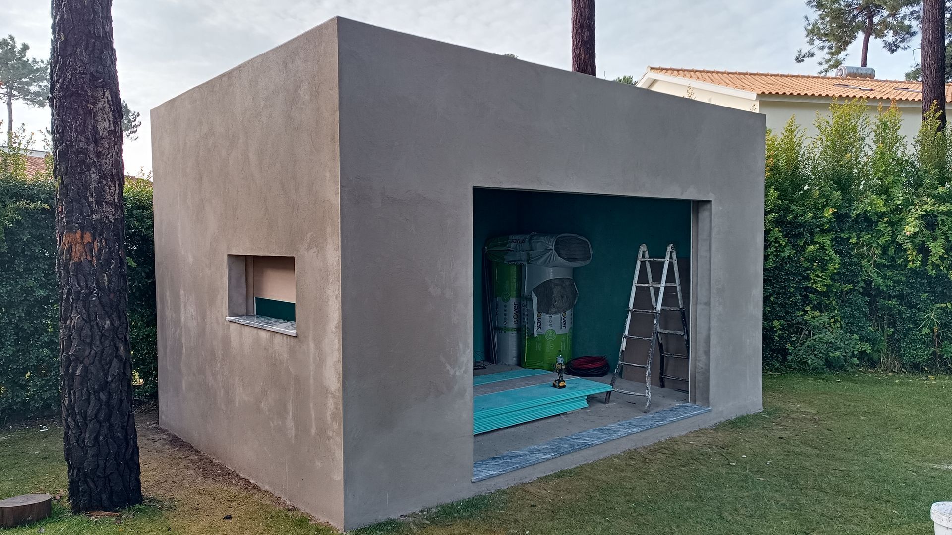 Exterior de um galpão de estuque cinza com uma abertura e uma pequena janela no quintal. Interior com parede verde-azulada e uma escada.