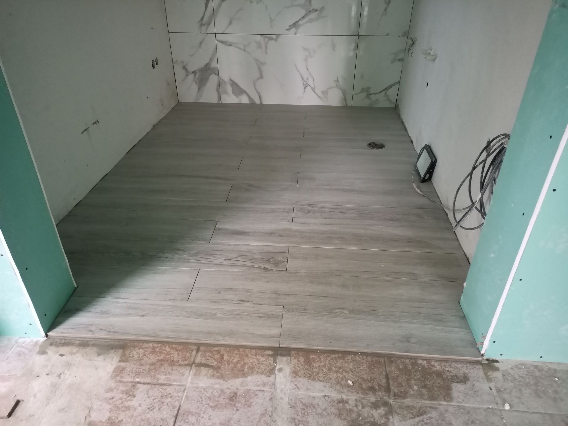 Banheiro em construção, com piso em ladrilhos imitando madeira cinza e azulejos nas paredes imitando mármore branco.