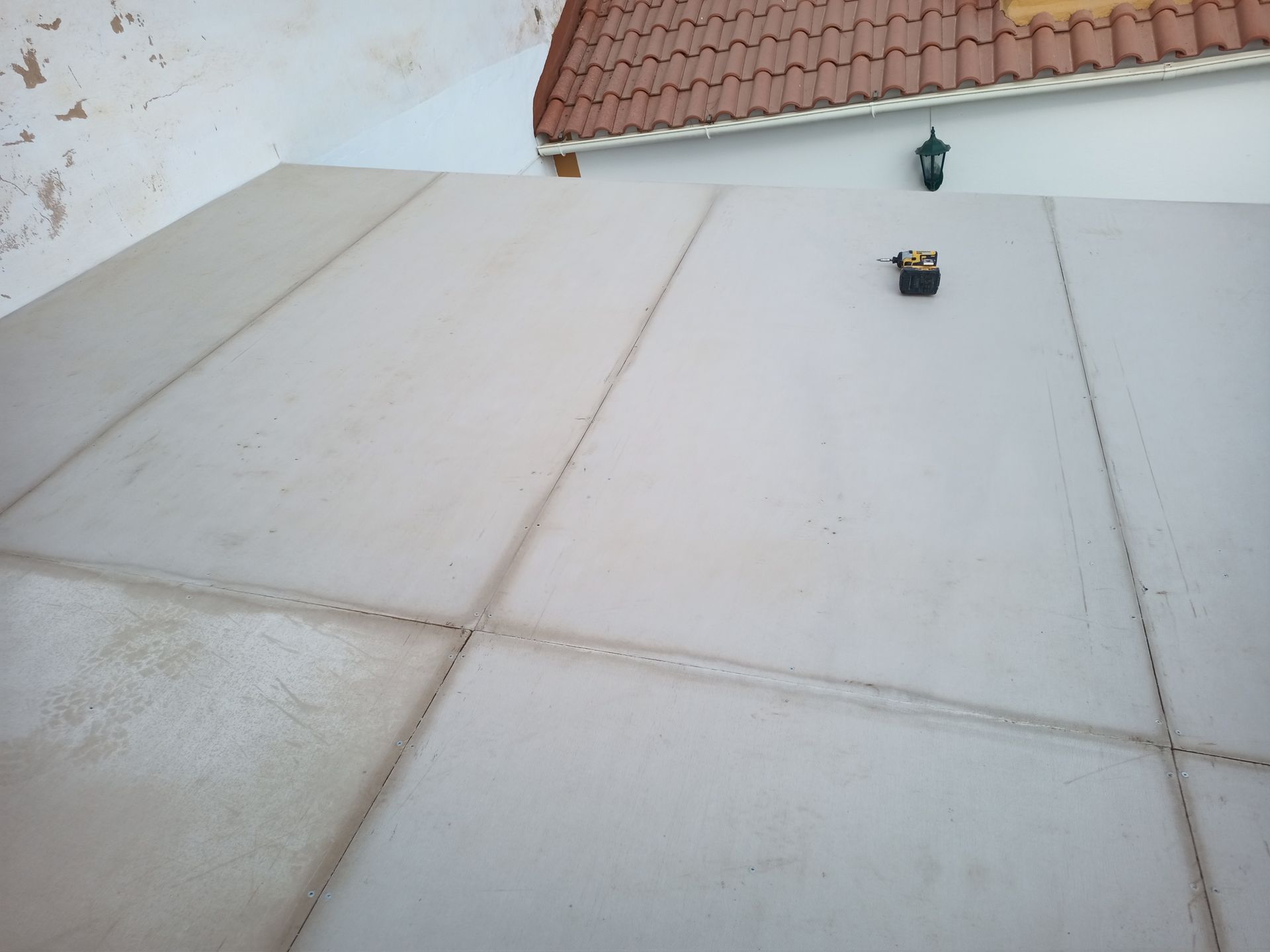 Parede de concreto cinza com linhas escuras, uma pequena caixa preta e um telhado de telha cerâmica.