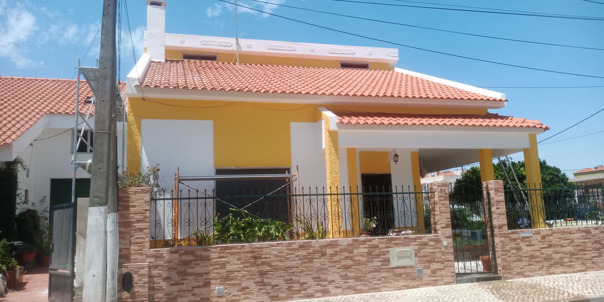 Casa amarela e branca com telhado de telha vermelha, muro de tijolos e portão de ferro forjado.