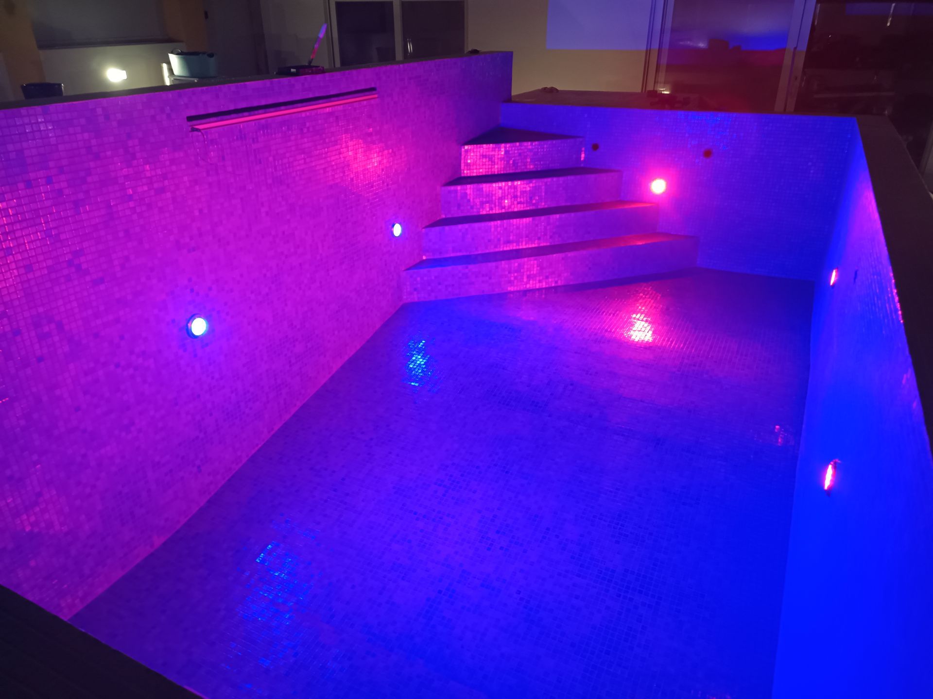 Piscina iluminada com azulejos em mosaico, com luzes roxas e azuis. Uma escada leva até a água.