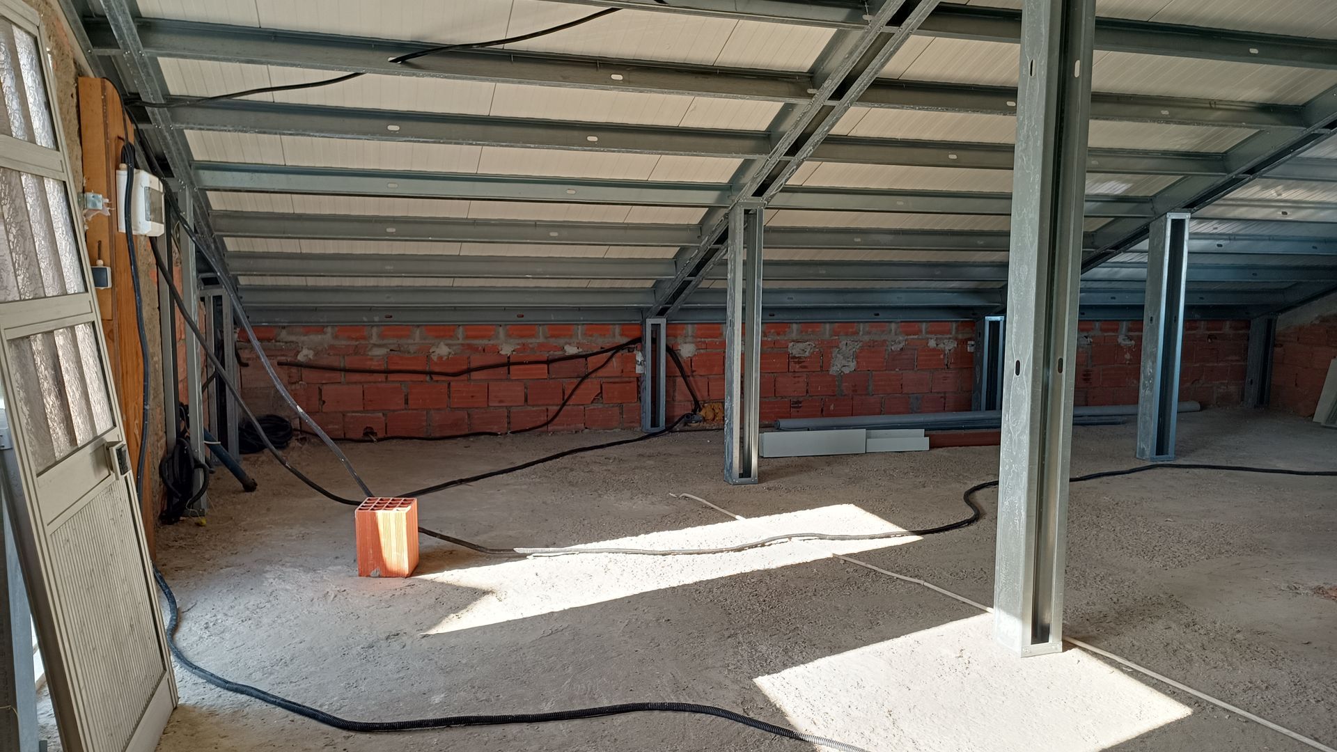 Vista interna de um canteiro de obras com vigas de aço expostas, paredes de tijolos vermelhos e fiação elétrica.