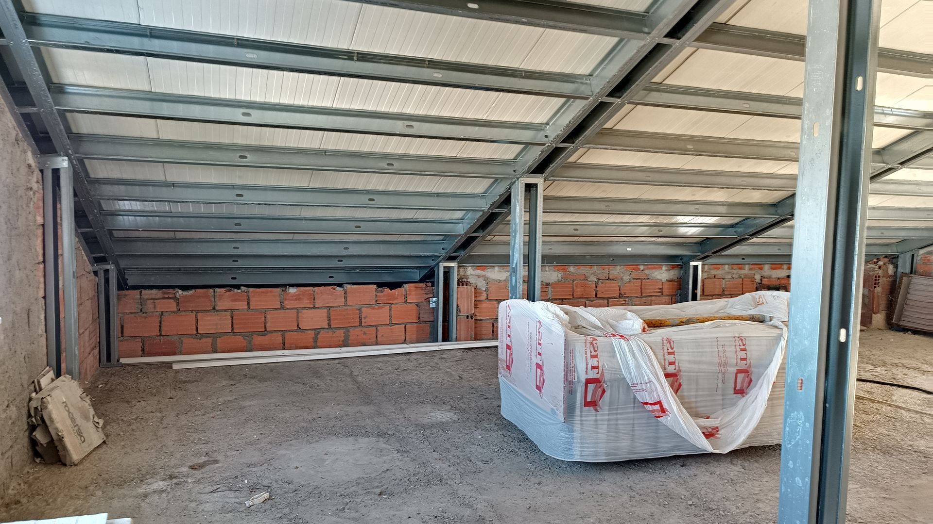 Vista interna de um edifício em construção, mostrando a estrutura metálica, as paredes de tijolo e o isolamento.