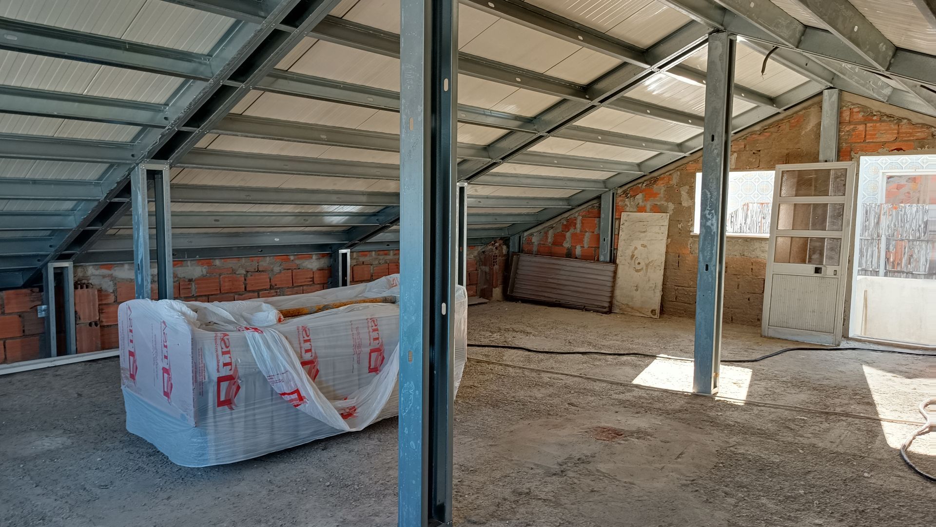 Interior do sótão inacabado com vigas de aço expostas, paredes de tijolo e isolamento.
