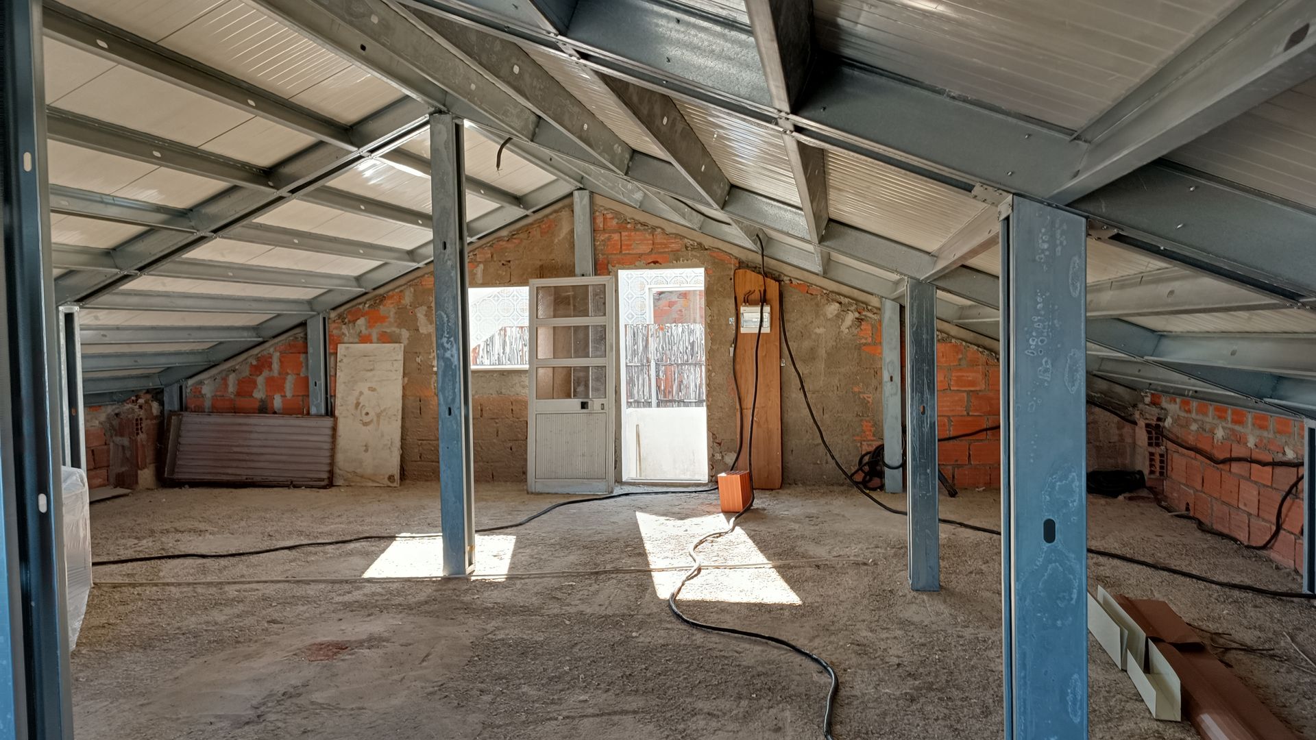 Interior de um canteiro de obras. Vigas de aço e paredes de tijolos aparentes. Telhado e piso inacabados.