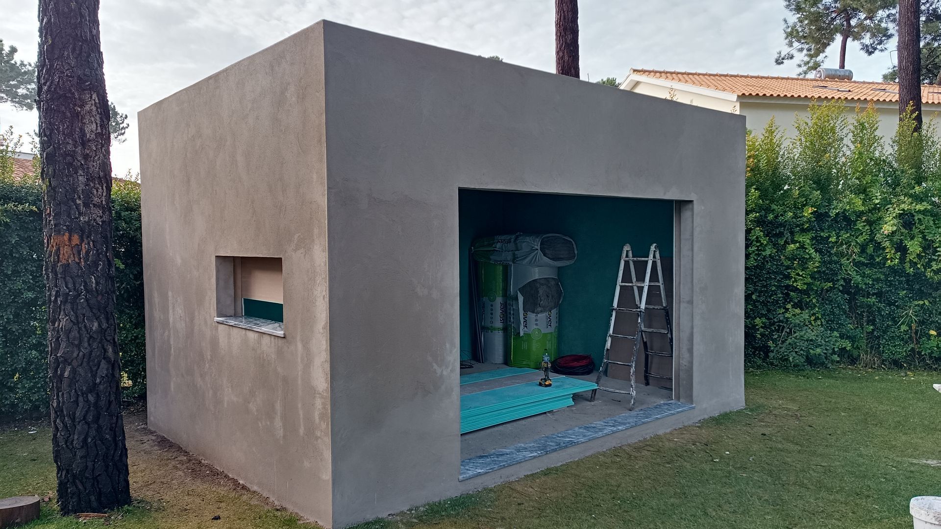 Estrutura cinza de cimento em forma de caixa, com a frente aberta e uma pequena janela quadrada, instalada ao ar livre na grama.