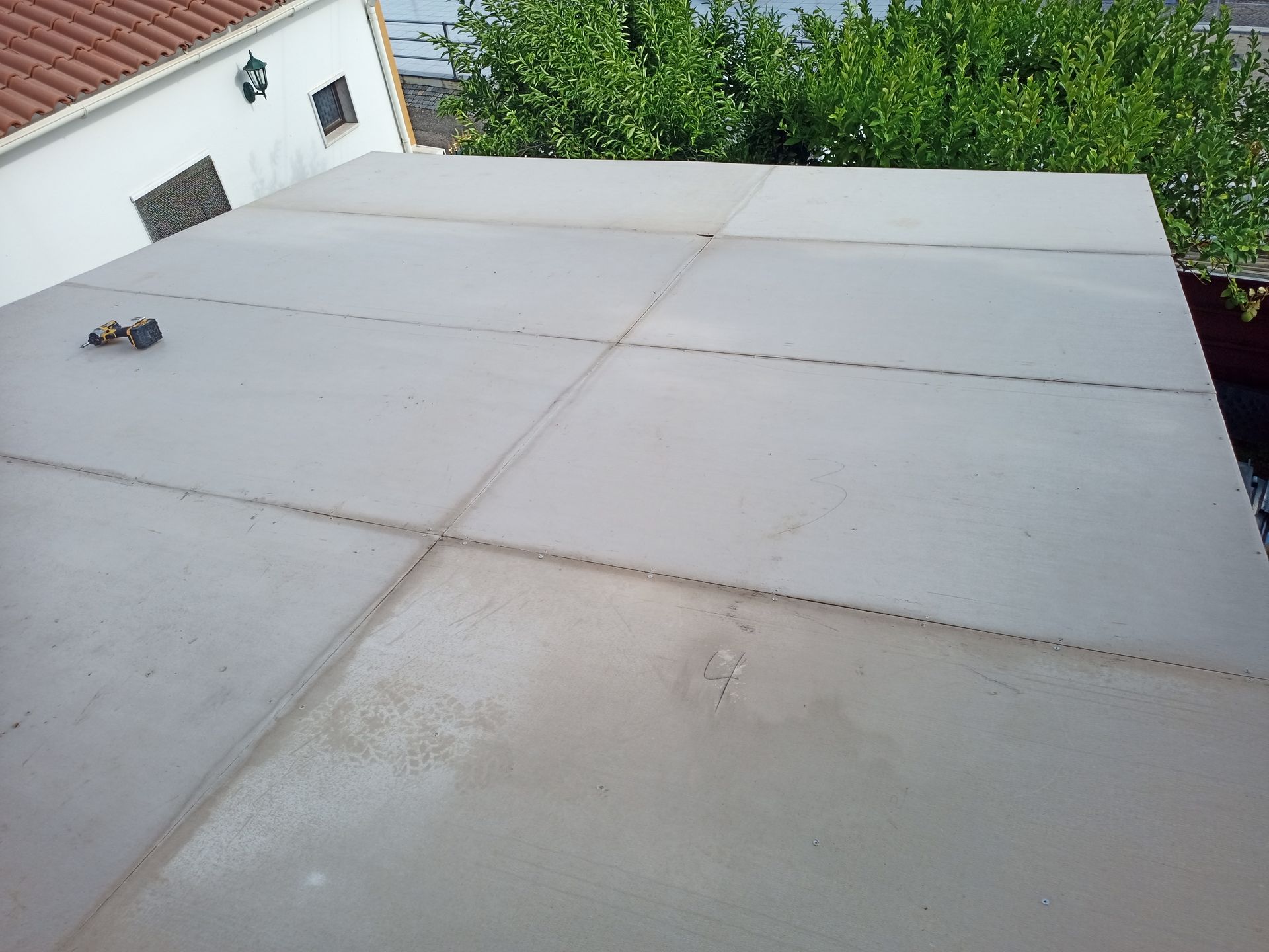 Telhado plano de telhas cinzentas com juntas visíveis. Folhagem verde e um edifício branco são visíveis ao fundo.