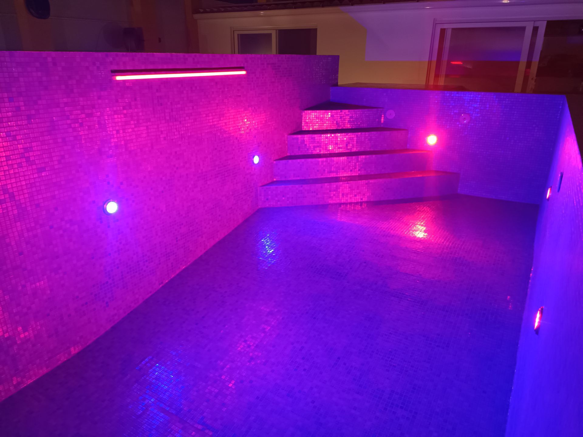 Piscina vazia iluminada de roxo, com degraus, azulejos em mosaico e luzes embutidas à noite.