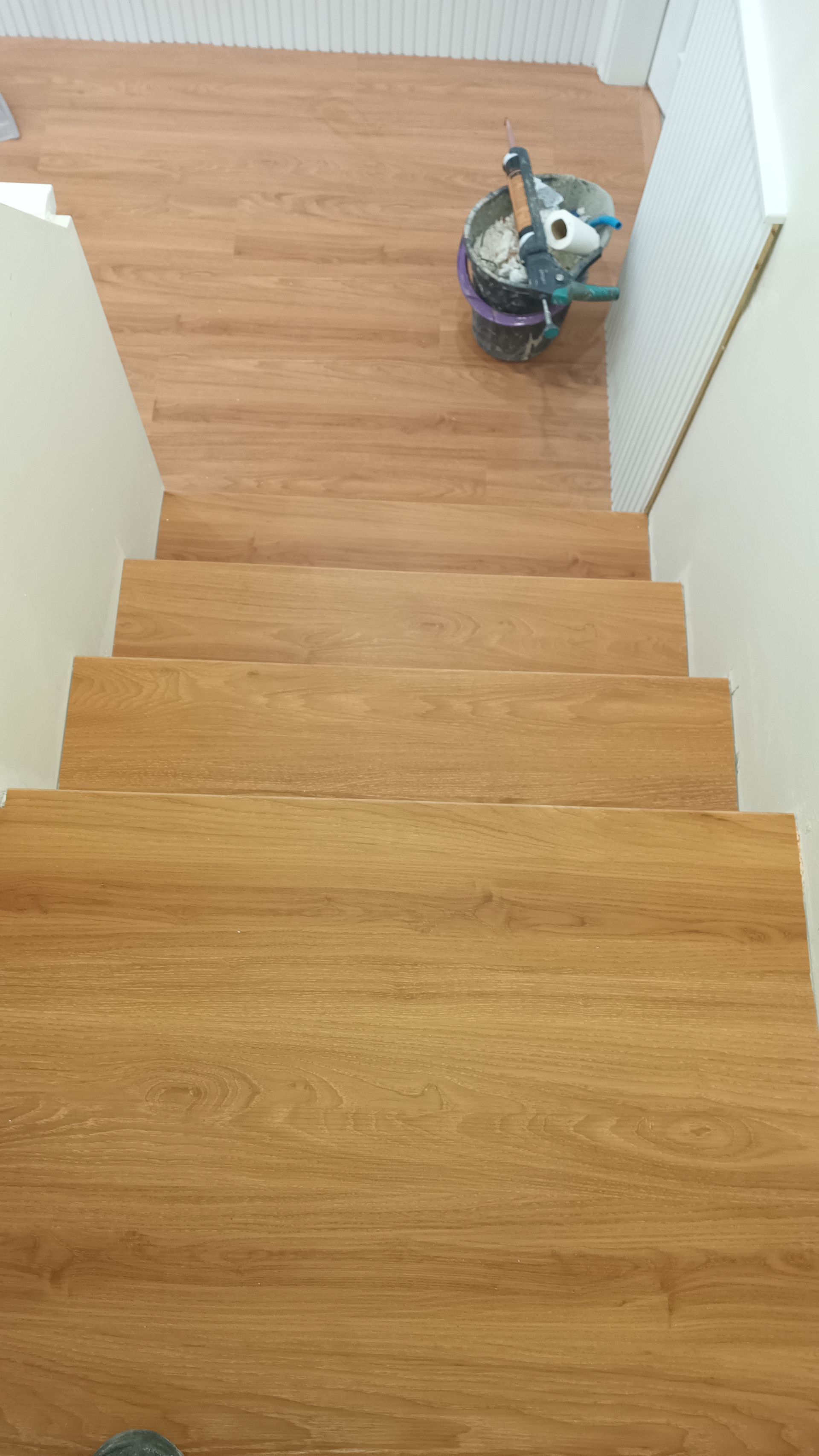 Escada de madeira que desce, de cor marrom claro. Há um balde no topo.