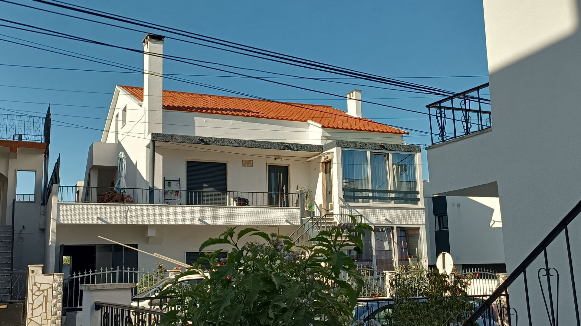 Casa branca com telhado laranja, varanda e fios elétricos. Arbustos verdes em primeiro plano.