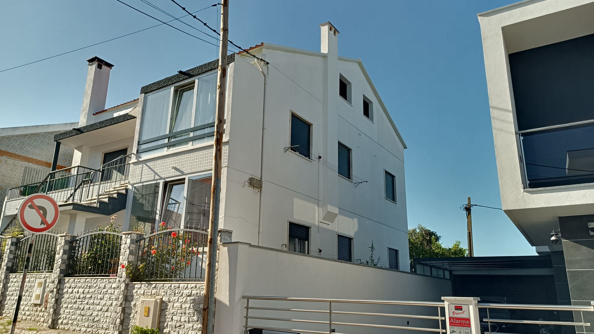 Prédio branco de três andares com janelas pequenas, uma chaminé e uma varanda envidraçada; céu azul claro ao fundo.