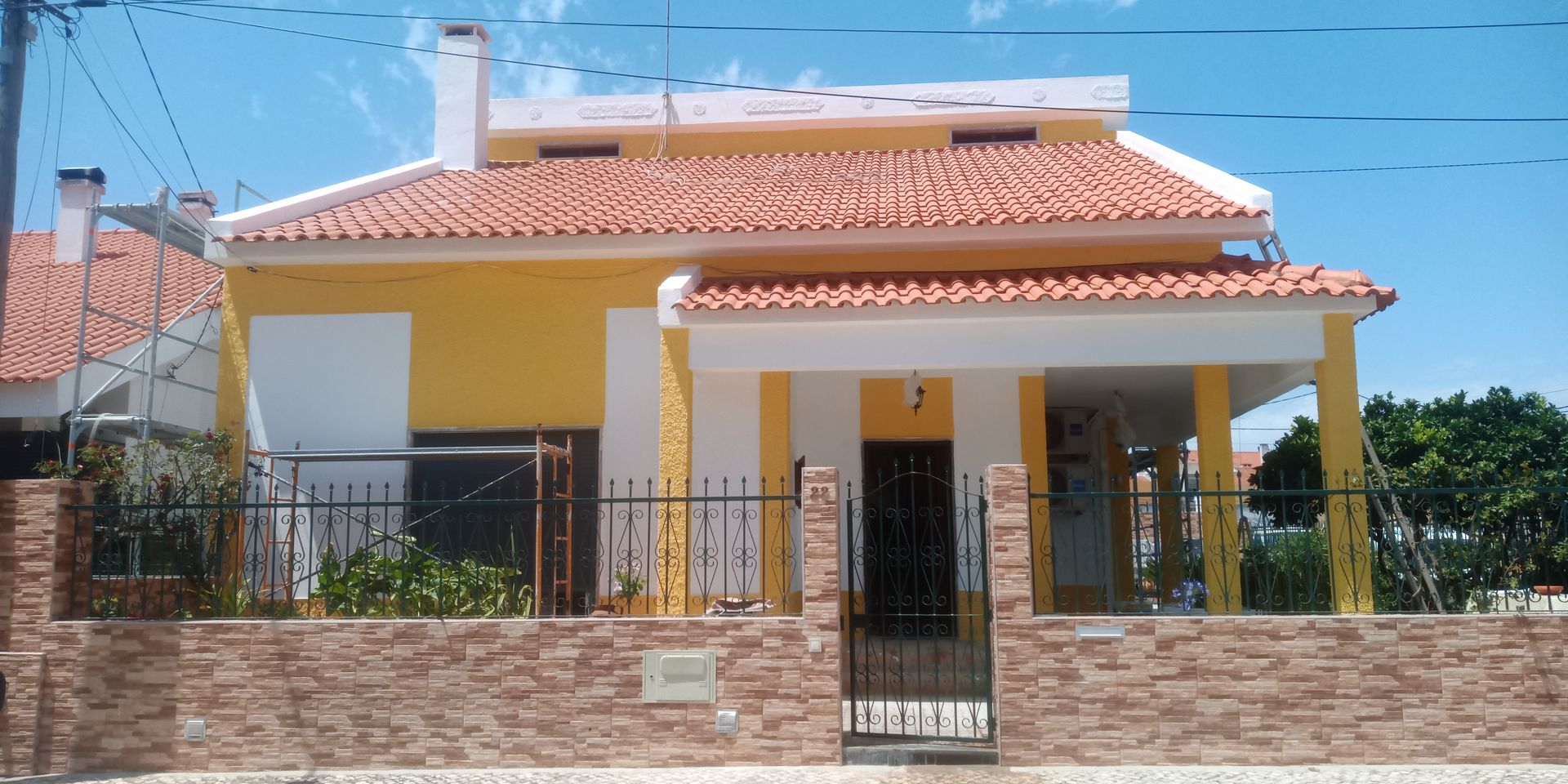 Casa amarela com telhado de telhas vermelhas, muro de tijolos e portão de ferro preto sob um céu azul claro.