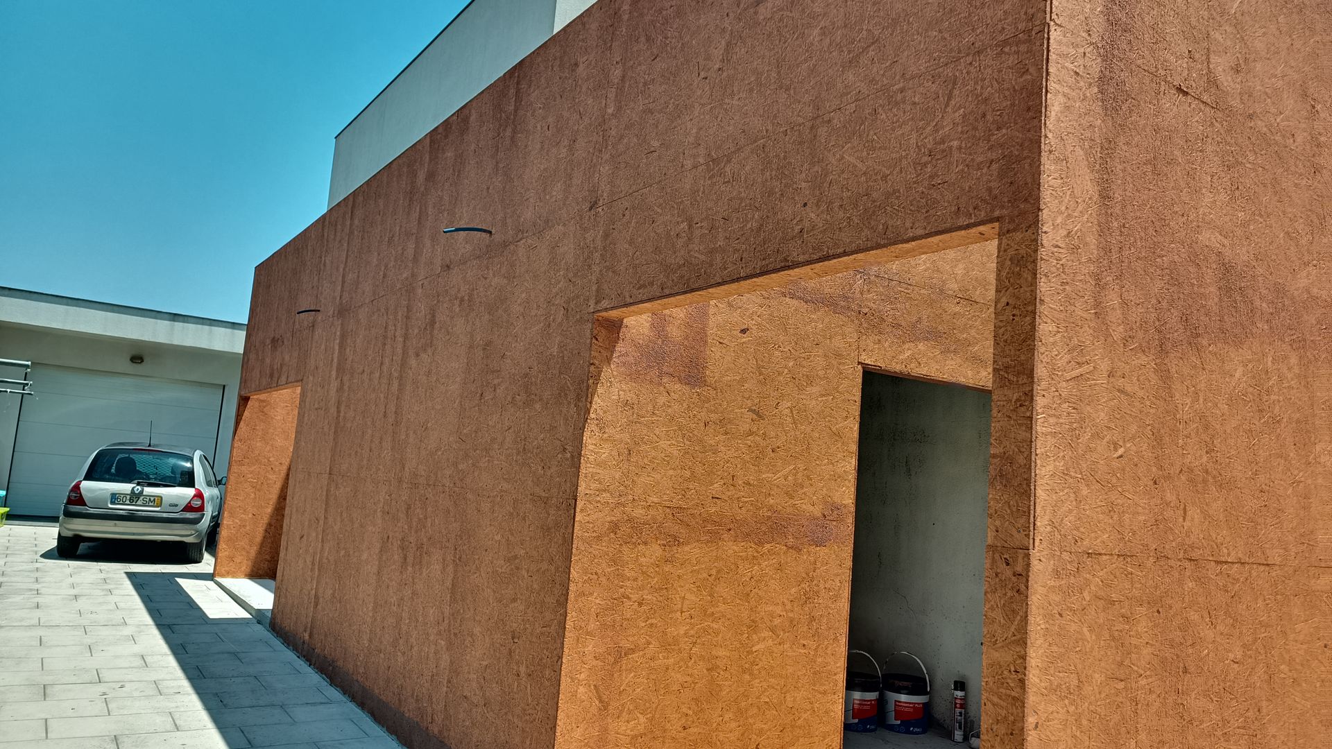 Exterior de um edifício com paredes texturizadas em tom marrom, porta de entrada e carro estacionado. Céu azul ao fundo.