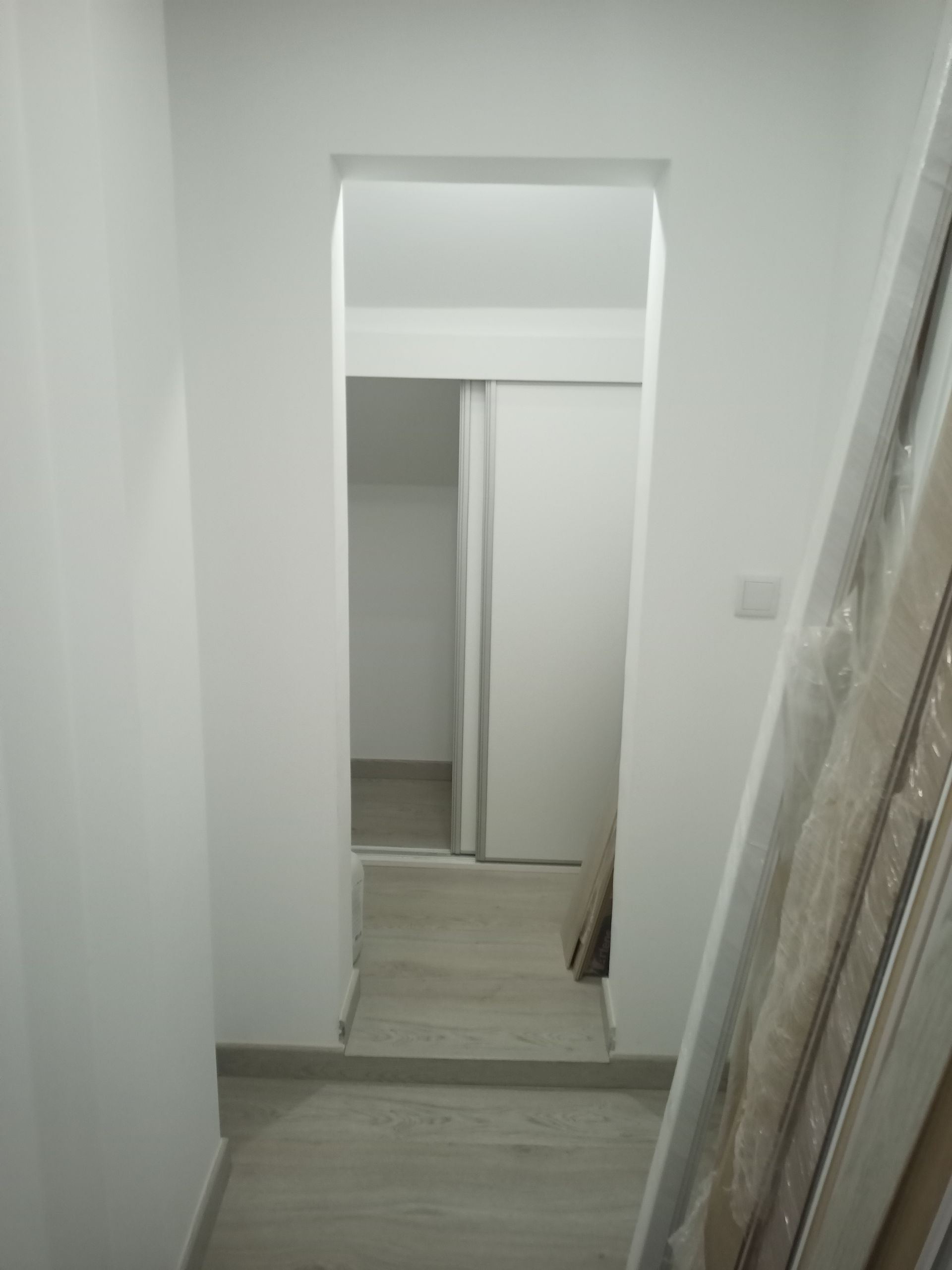 Corredor branco com porta em arco que dá acesso a uma sala com portas de correr fechadas e piso cinza claro.