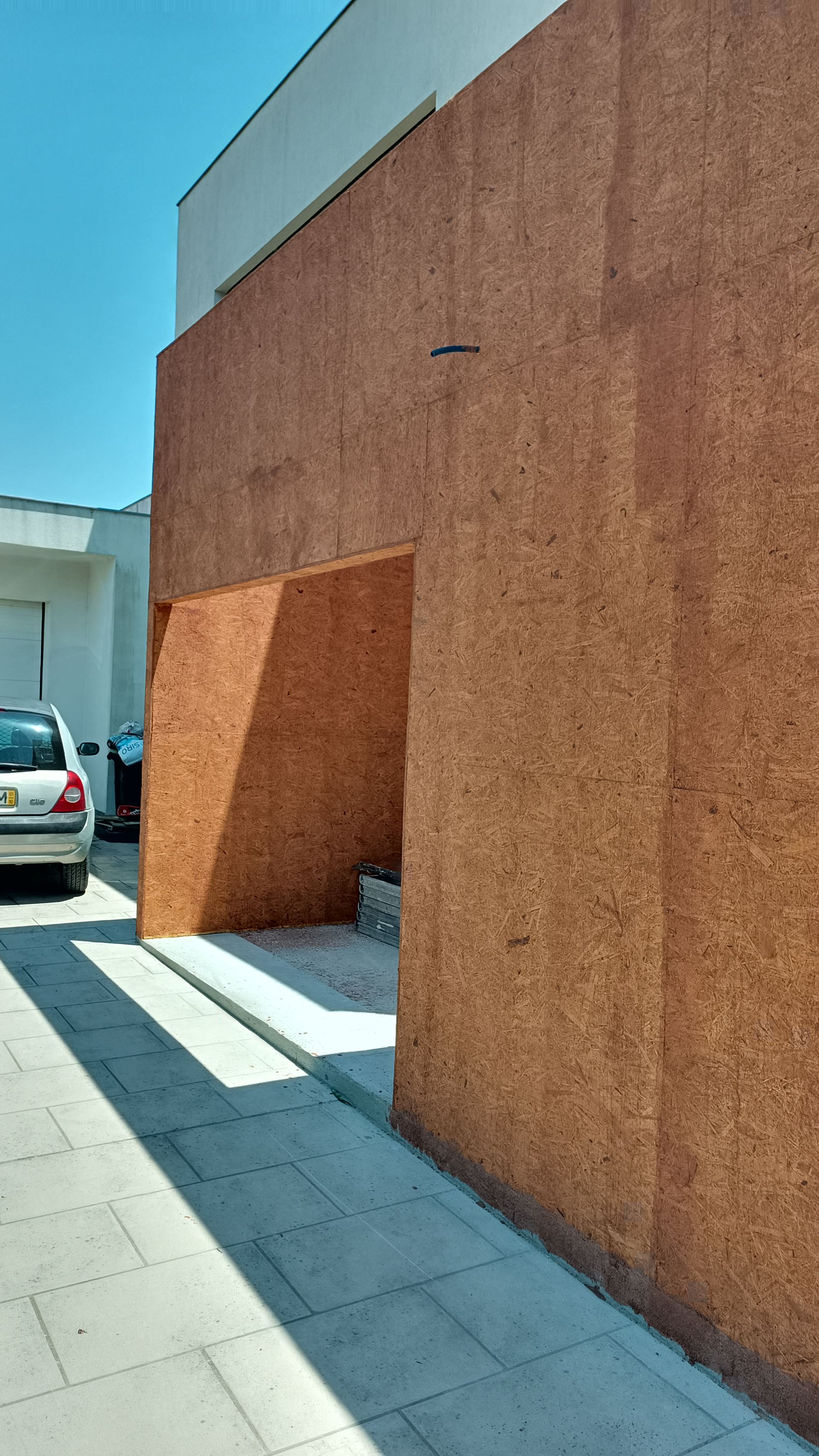 Fachada de edifício com textura marrom e uma porta de entrada; um carro está estacionado ao fundo.
