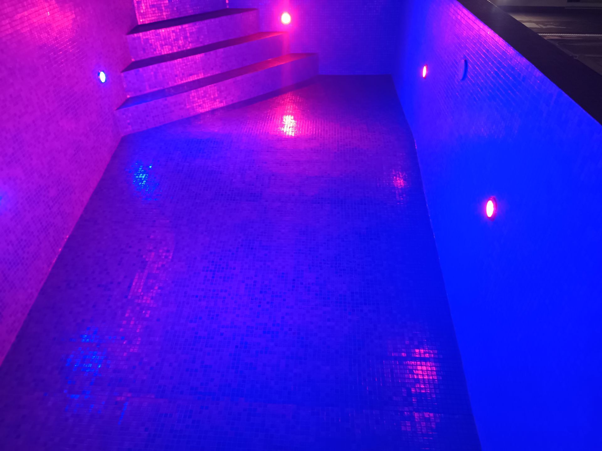 Piscina com azulejos azuis no interior, degraus e luzes rosa e azuis refletindo na água.