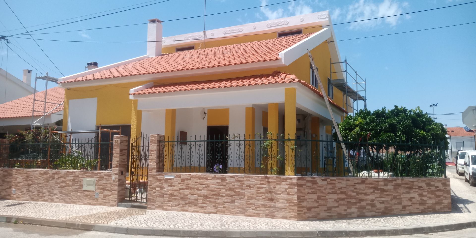 Casa amarela com telhado de telha vermelha, detalhes brancos, parede de tijolos e cerca de ferro forjado sob um céu azul.