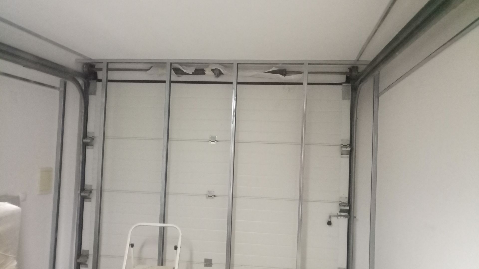 Vista interna da estrutura da porta de um reboque de carga, com suportes metálicos expostos e uma porta branca parcialmente aberta.