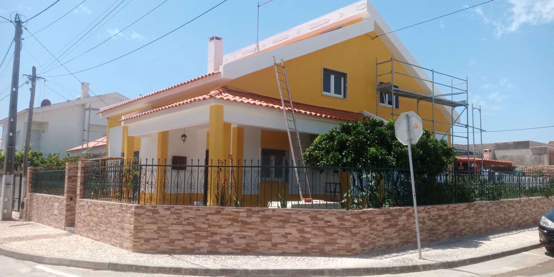 Casa amarela com detalhes brancos, telhas laranja e parede de tijolos. Há um andaime montado ao lado.
