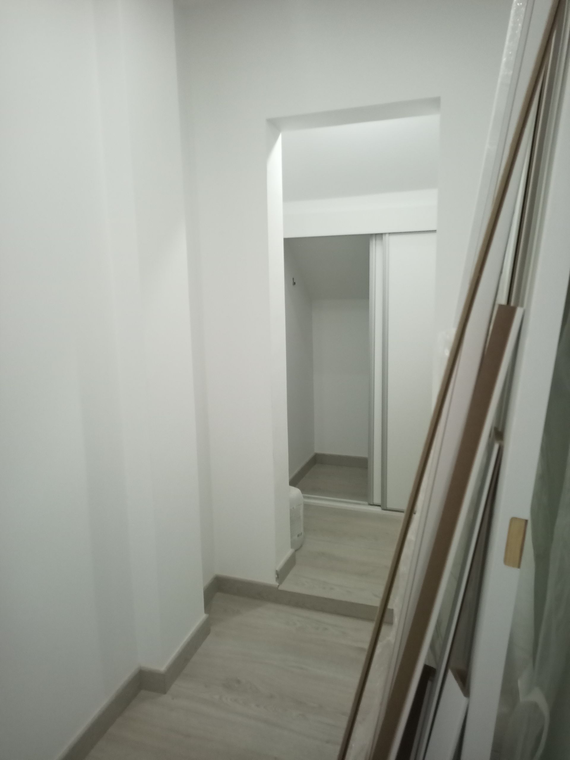 Corredor estreito com paredes brancas e piso que imita madeira clara. A porta dá acesso a um pequeno espaço de armazenamento. Tábuas encostadas na parede.
